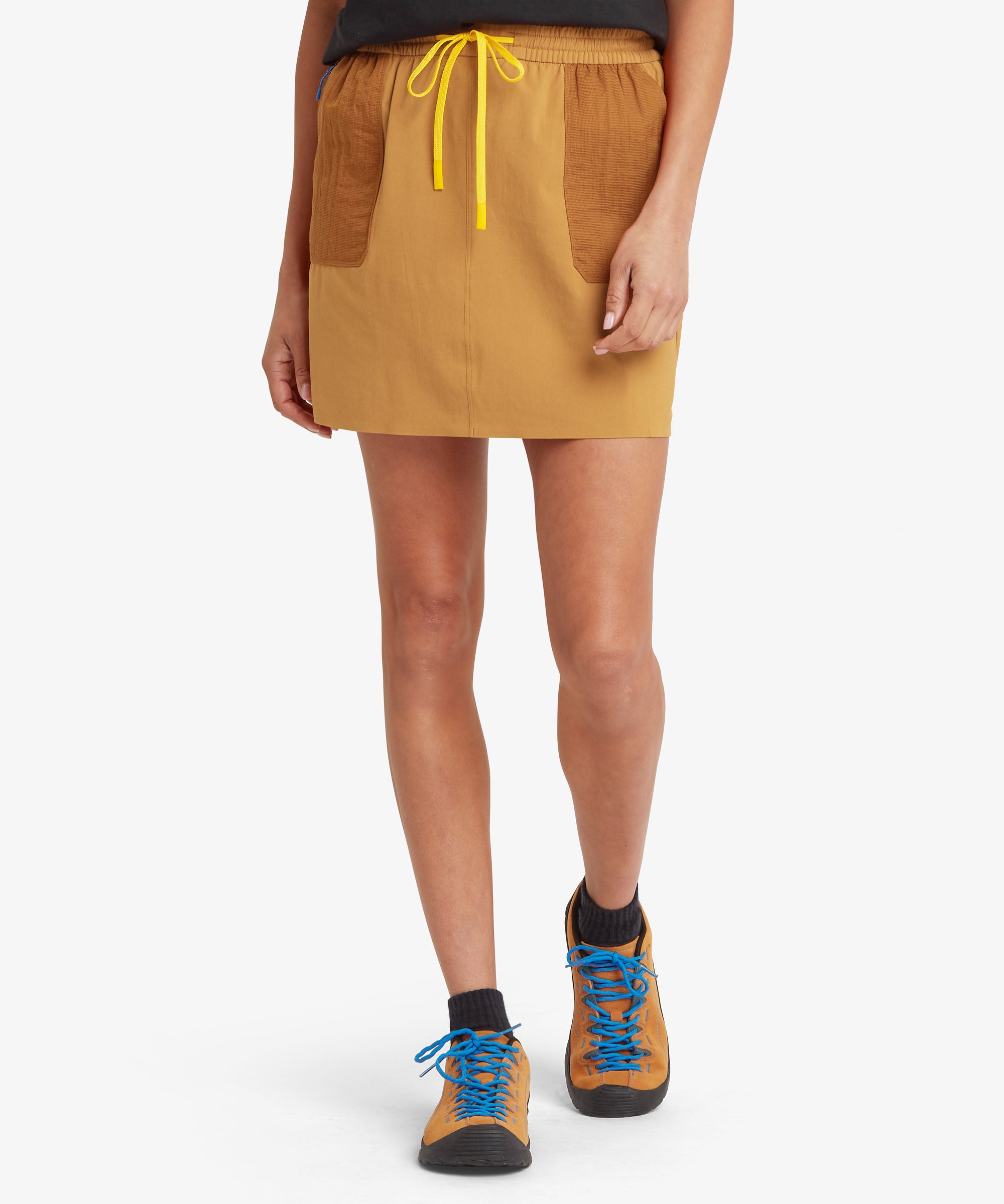 Full-body view of mustard-yellow skort with contrasting pockets and yellow drawstring, paired with outdoor shoes.