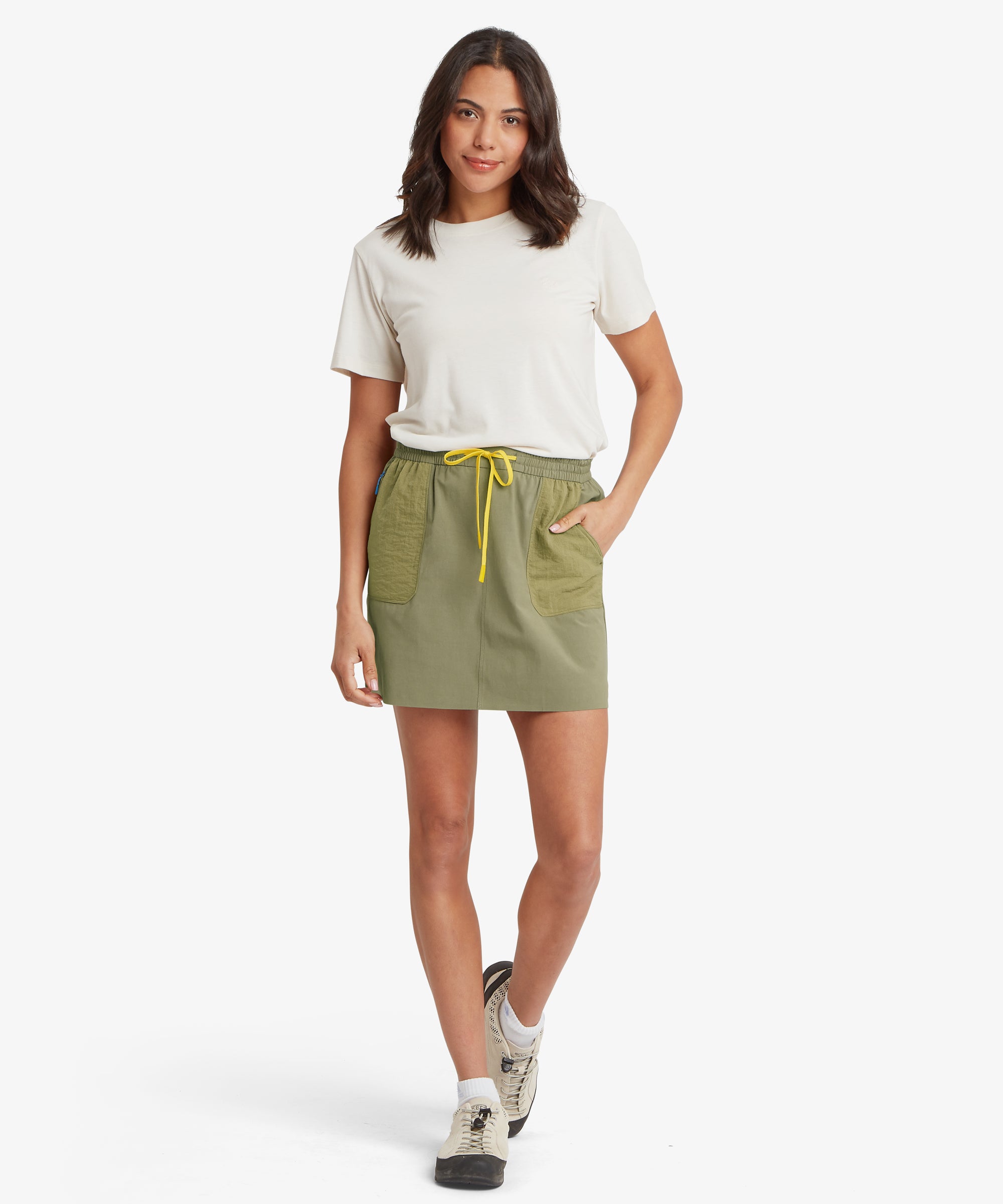 A woman wearing an olive green drawstring skort with a relaxed, above-the-knee fit that sits at the natural waist, paired with a white short-sleeve T-shirt and casual sneakers.