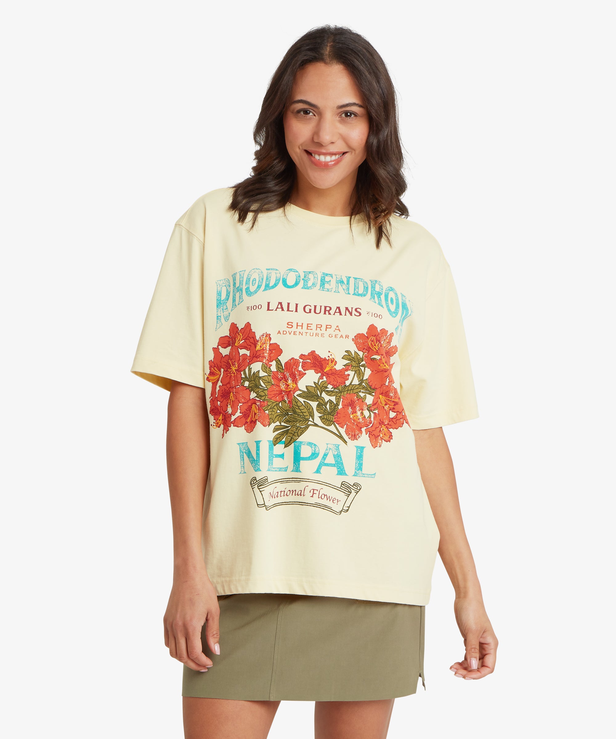 Eine Frau trŠgt ein gelbes T-Shirt mit Grafik, einer lockeren, entspannten Passform und Ÿberschnittenen Schultern, das mit einer auffŠlligen, von Nepal inspirierten Blumengrafik in Rot mit tŸrkisfarbener und blauer Schrift verziert ist, dazu einen olivgrŸnen Rock.