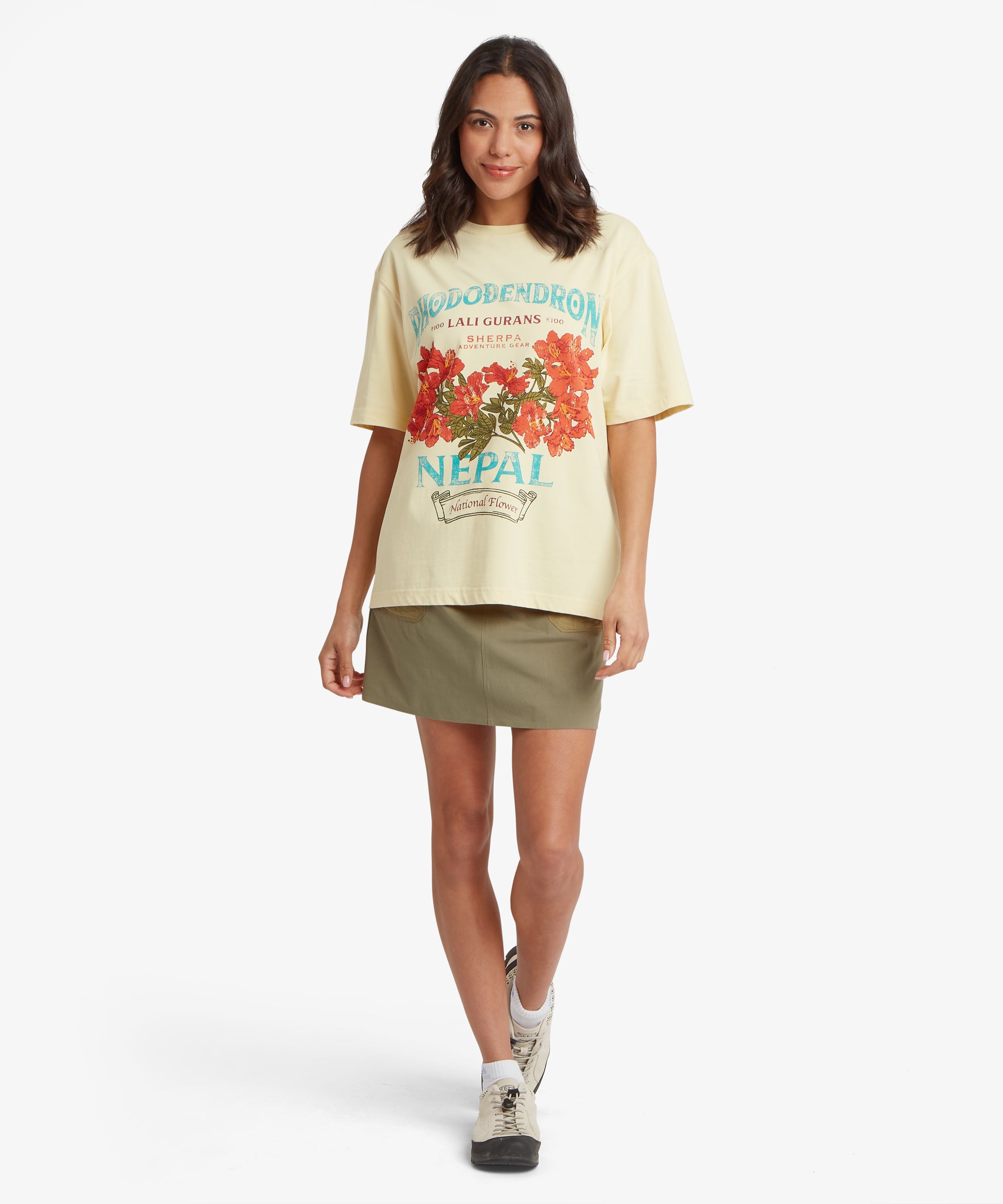 Frau, die auf die Kamera zugeht und ein gelbes T-Shirt mit Grafik trŠgt, das locker und oversized geschnitten ist und auf der Vorderseite eine gro§e rote Blumengrafik und tŸrkisfarbene SchriftzŸge aufweist.