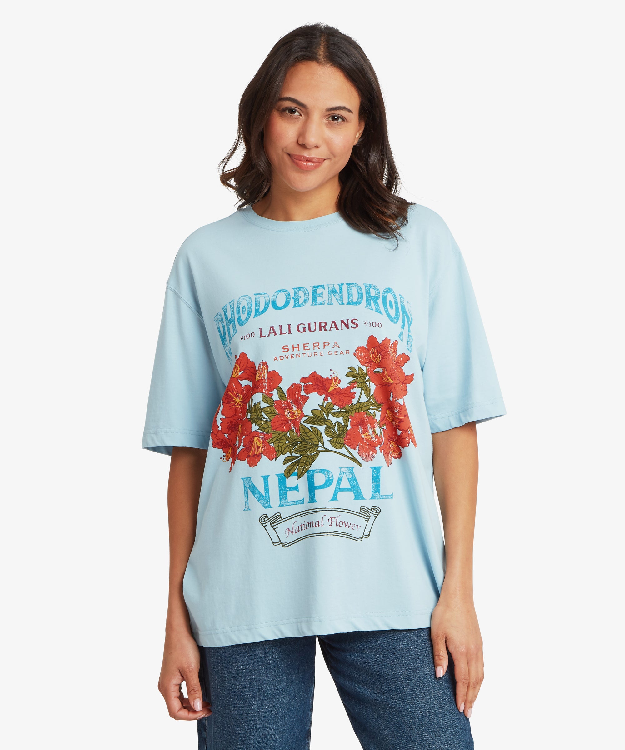 Frau in einem hellblauen T-Shirt mit Grafikprint, lockerer, entspannter Passform und leicht Ÿberschnittenen Schultern, mit auffŠlligem rotem Blumenmuster im Nepal-Stil und tŸrkis- und marineblauer Schrift, kombiniert mit einer blauen Jeans.