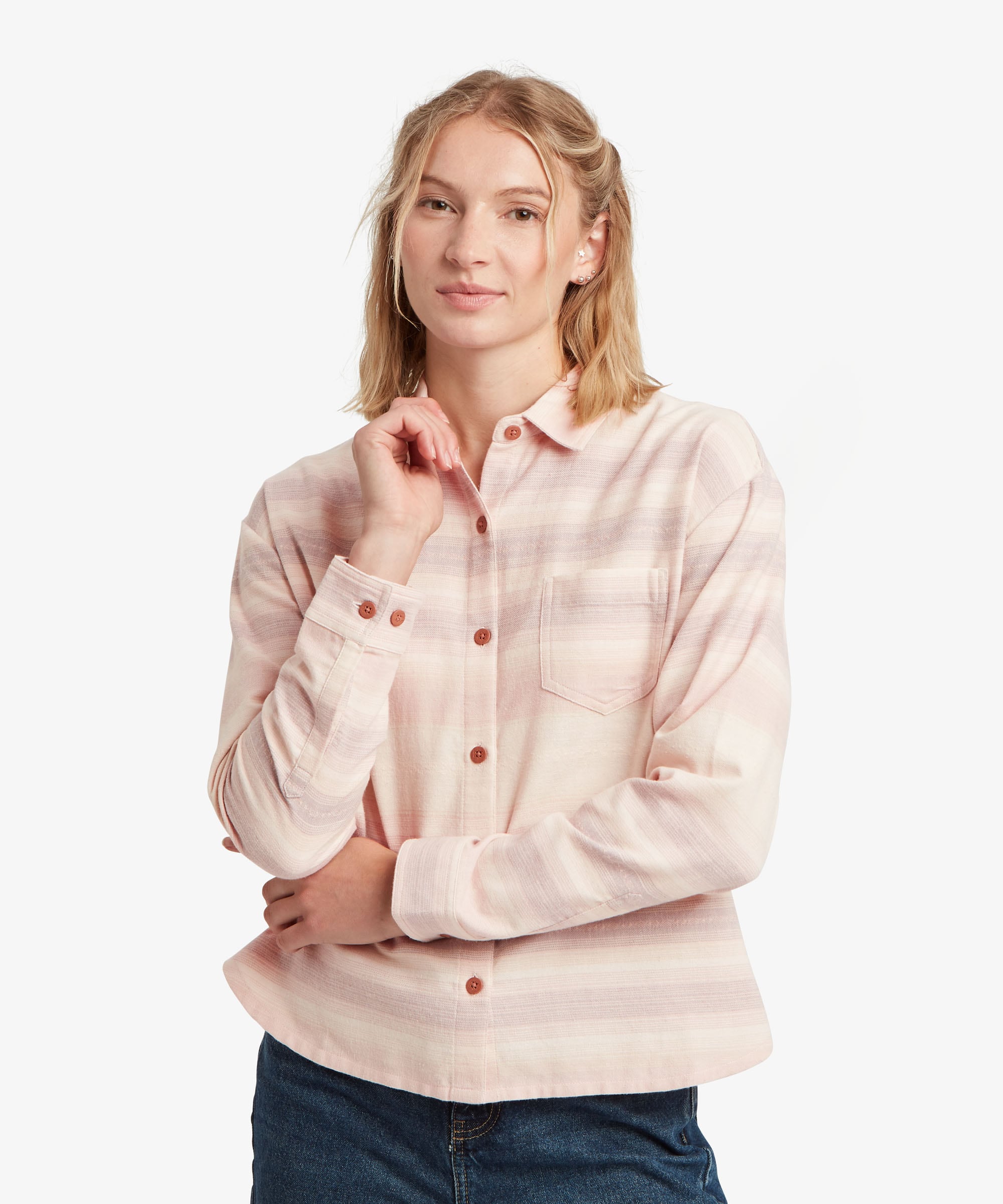A woman modeling a light pink and white striped long-sleeve button-up shirt with brown buttons, paired with dark blue jeans, against a plain white background.