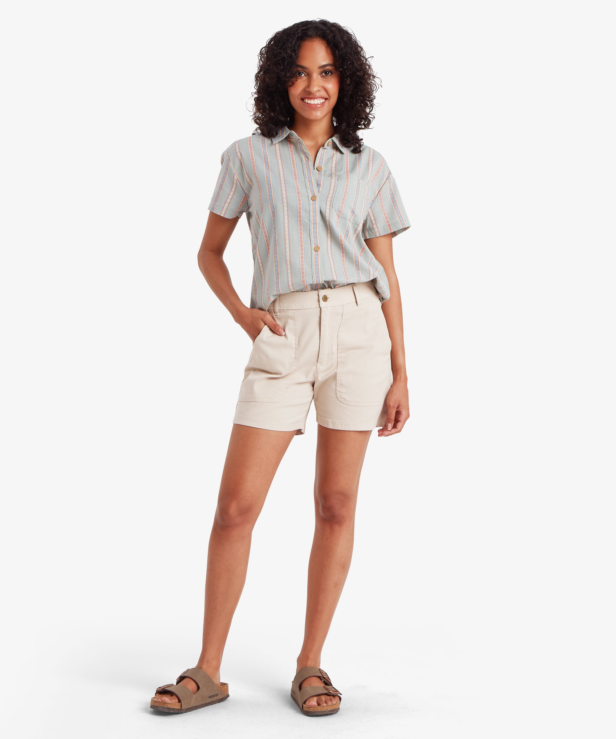 A full-length view of a woman walking towards the camera wearing beige utility shorts and a striped short-sleeved shirt. The shorts have a straight, relaxed silhouette that falls mid-thigh, offering a comfortable and airy fit for outdoor activities.
