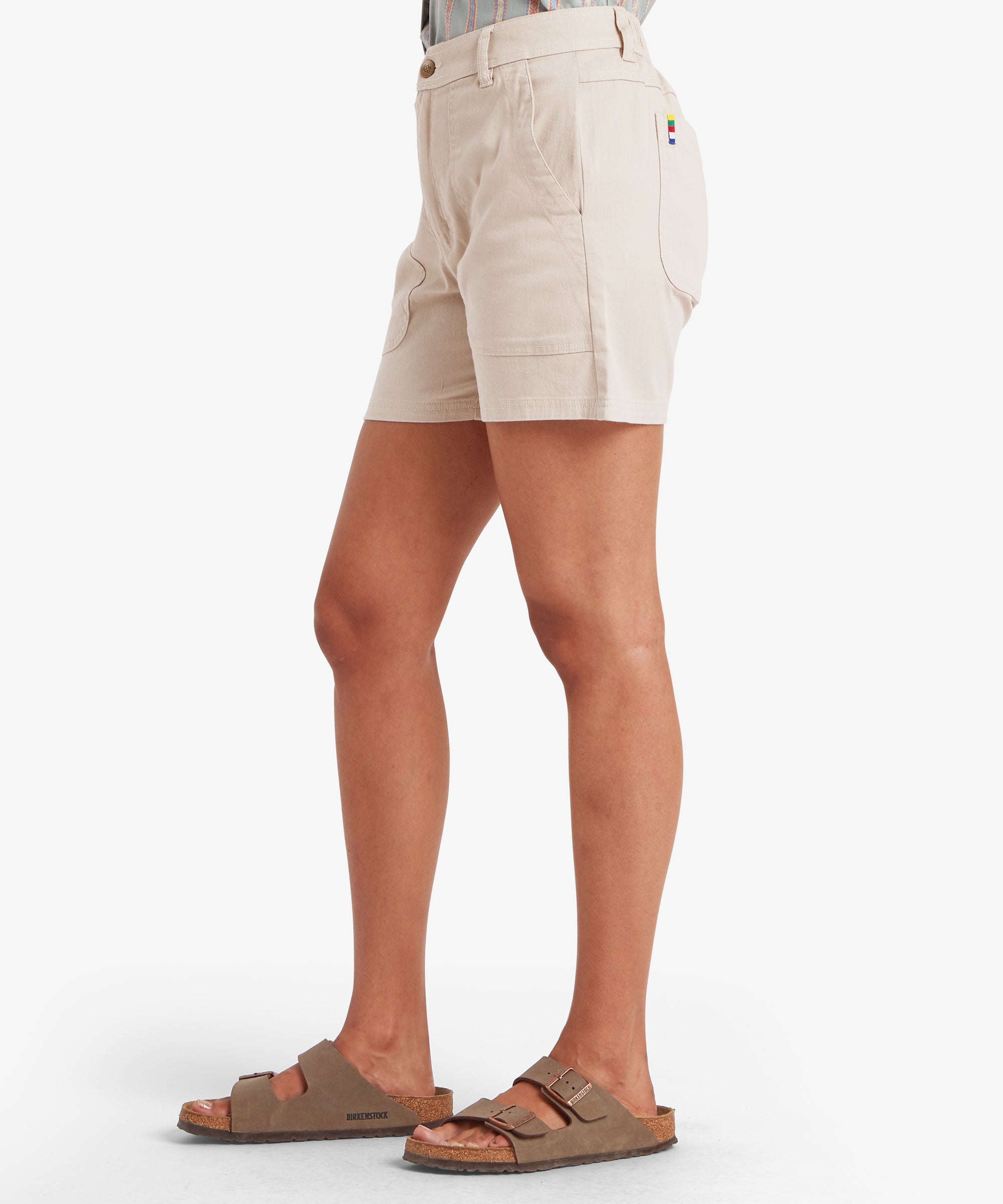 A profile view of the beige utility shorts, highlighting the deep side pockets and the clean, straight-cut hem. The silhouette is loose through the hips and thighs, paired with brown two-strap sandals for a functional, travel-ready style.