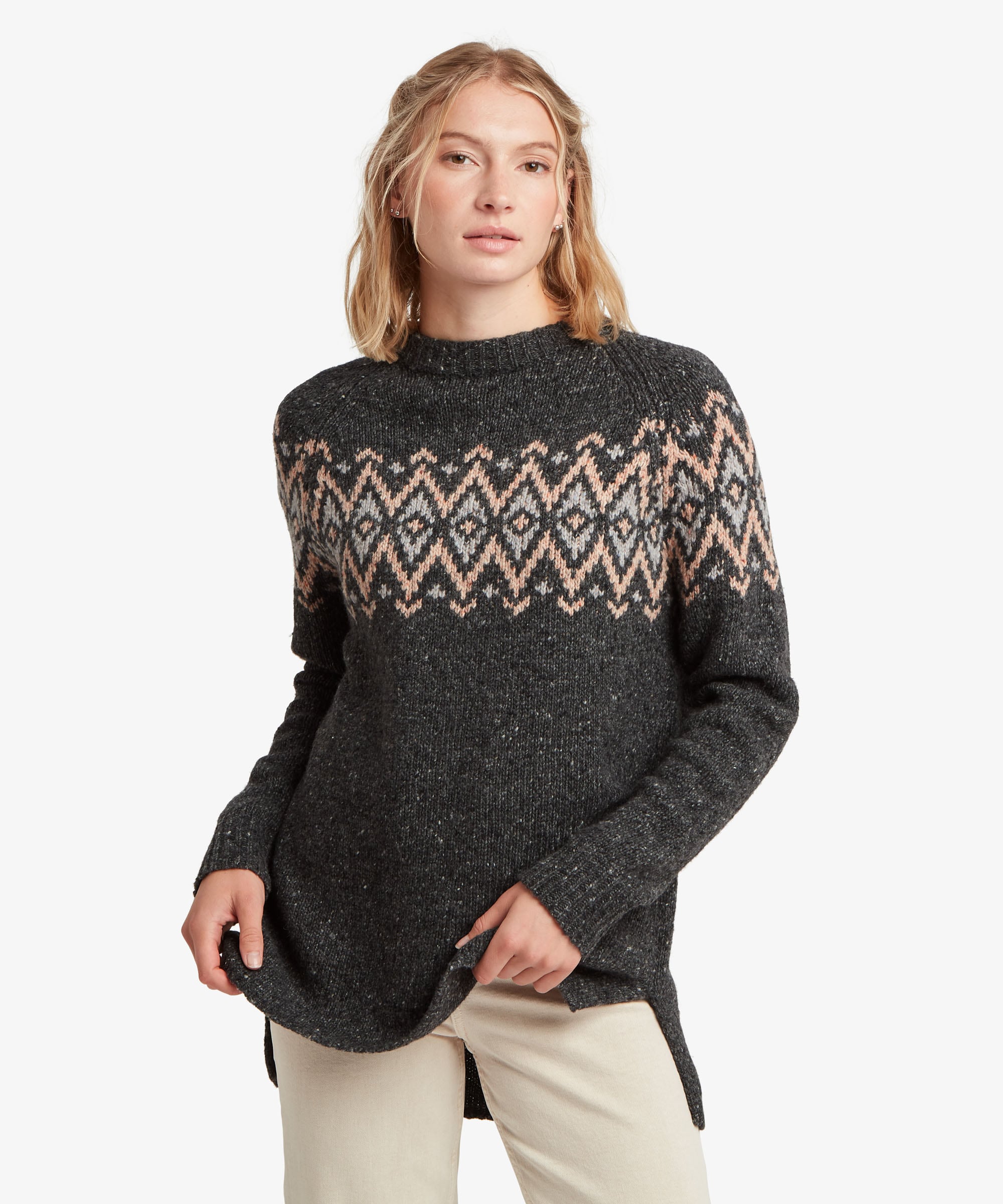 A female model standing against a plain white studio background wearing the Sherpa Adventure Gear Dumji Long Sweater in black, shown from the front. The long knitted sweater features a relaxed silhouette, long sleeves and a ribbed neckline, with a contrasting geometric patterned band running across the upper chest and continuing around the sleeves. She wears it with light-coloured trousers.