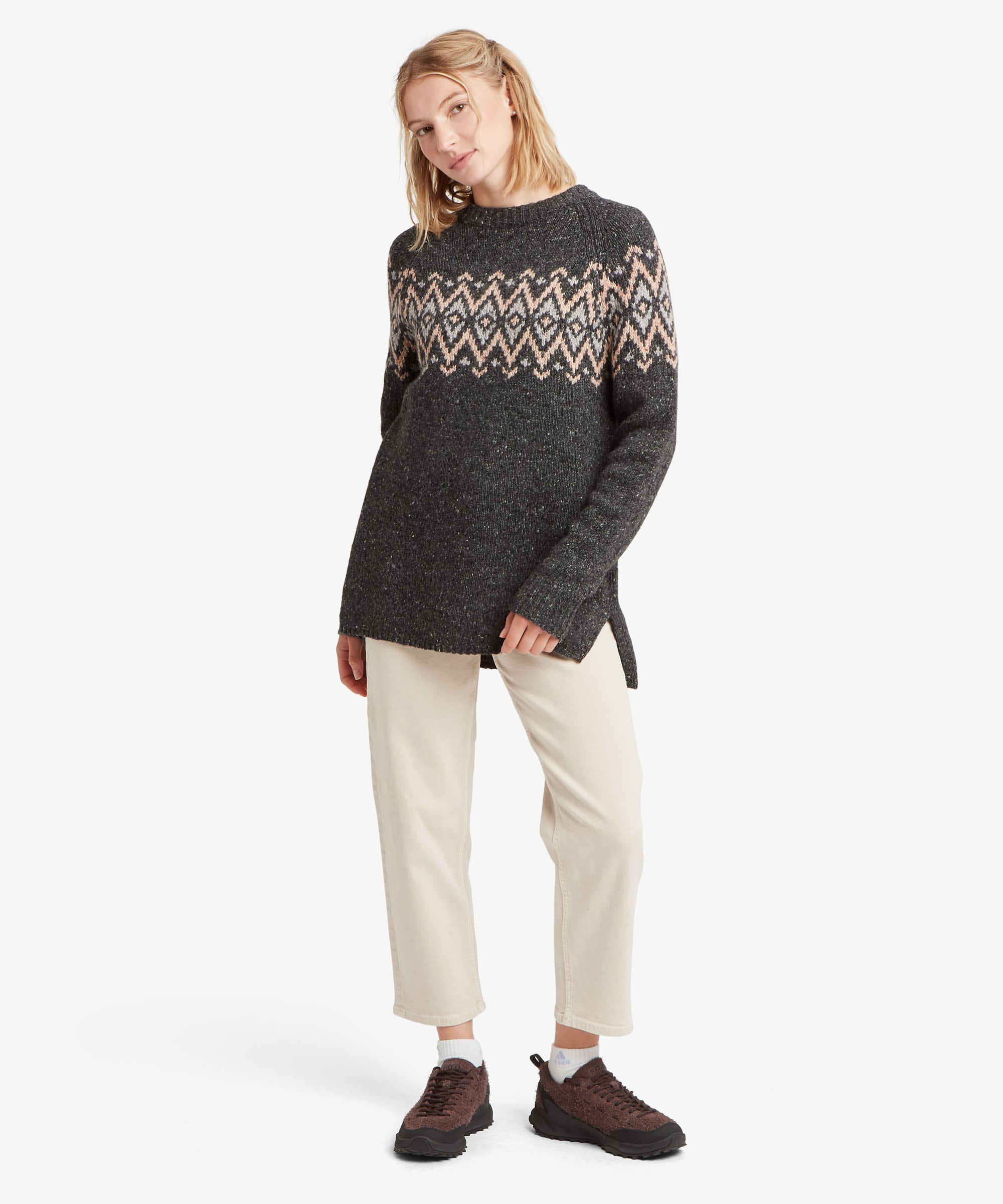 A female model, shown in full length, wearing the Sherpa Adventure Gear Dumji Long Sweater in black, shown from the front. The sweater has a longer silhouette extending over the hips, long sleeves and a ribbed crew neckline, with an intricate cream and beige geometric pattern across the upper chest and sleeves. The model wears it with light-coloured trousers and dark casual shoes.