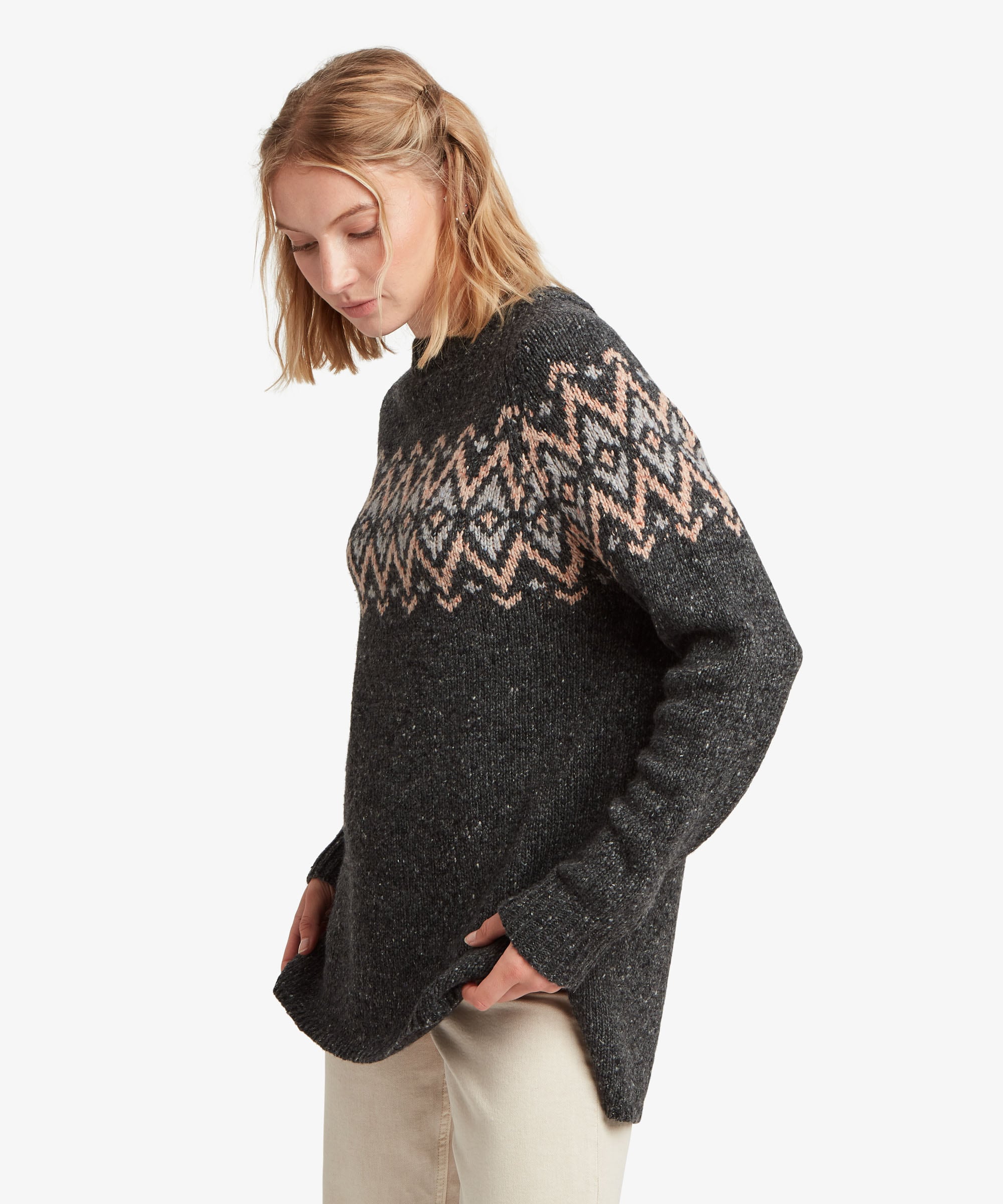 A female model, shown from the side, wearing the Sherpa Adventure Gear Dumji Long Sweater in black against a white studio background. The image shows the longer relaxed silhouette, long sleeves and ribbed neckline, with the contrasting geometric patterned band visible across the upper body and sleeve. The sweater extends over the hips for additional coverage.