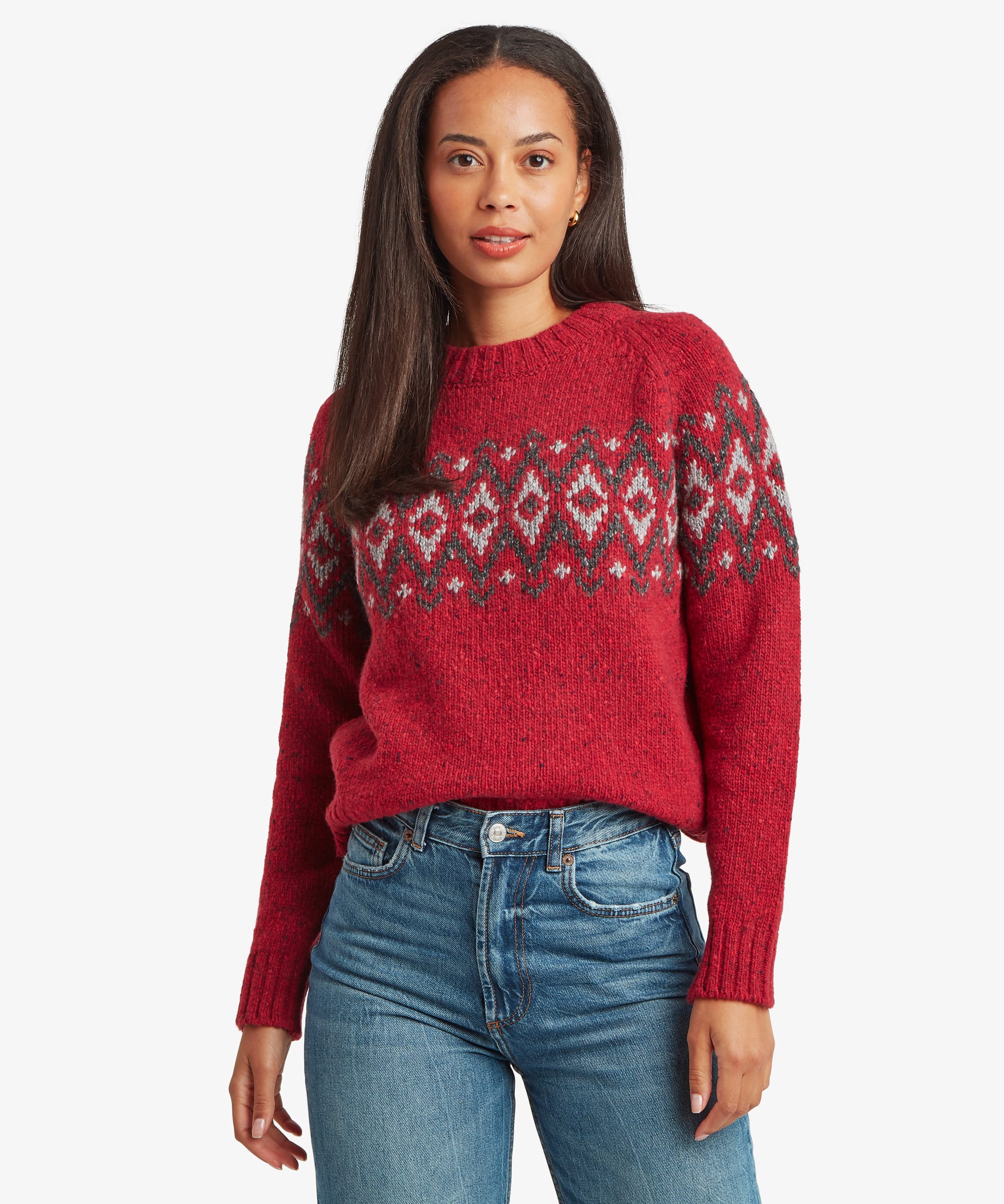 Woman wearing a bright red Fair Isle sweater with grey, white, and black geometric patterning across the chest and shoulders. Front view showing the crew neck and classic knit silhouette.