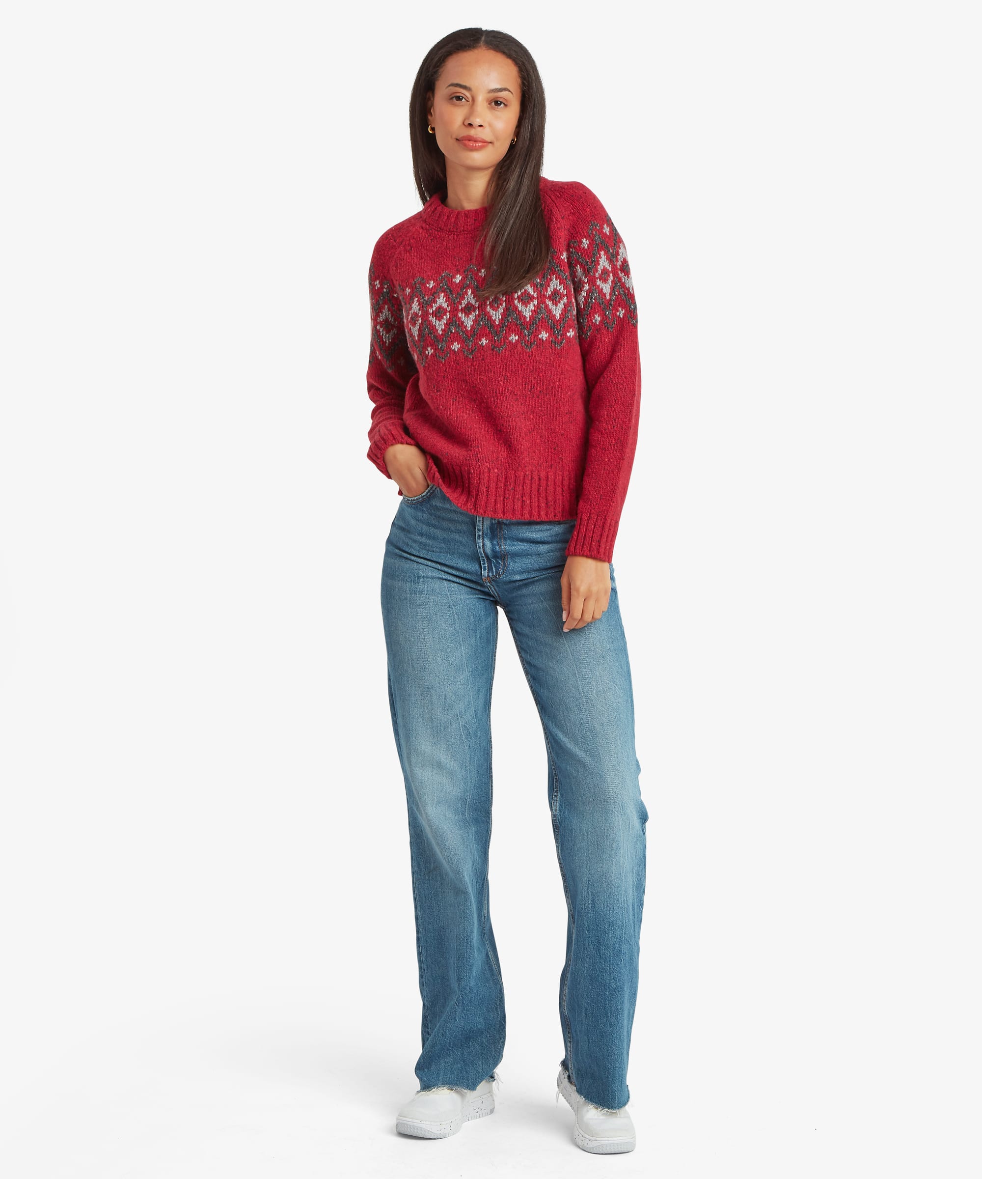 Full-body front view of a woman modeling a red Fair Isle sweater with a traditional Nordic-inspired yoke pattern. Styled with blue jeans for a casual outdoor look.