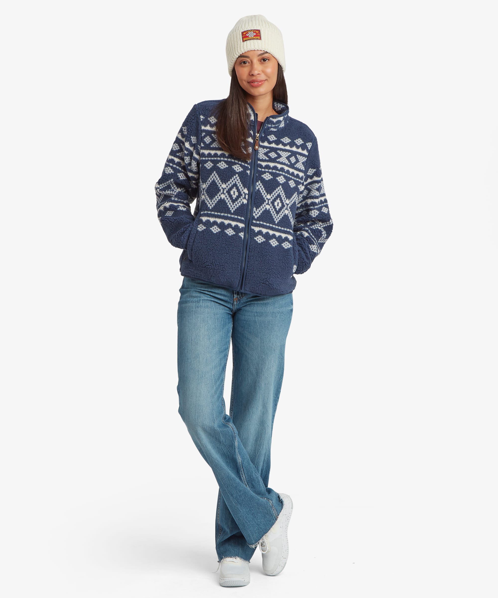 Full-body front view of a woman wearing a blue patterned fleece jacket and knit beanie. The jacket features a zip-front design, high collar, and soft sherpa-style texture.