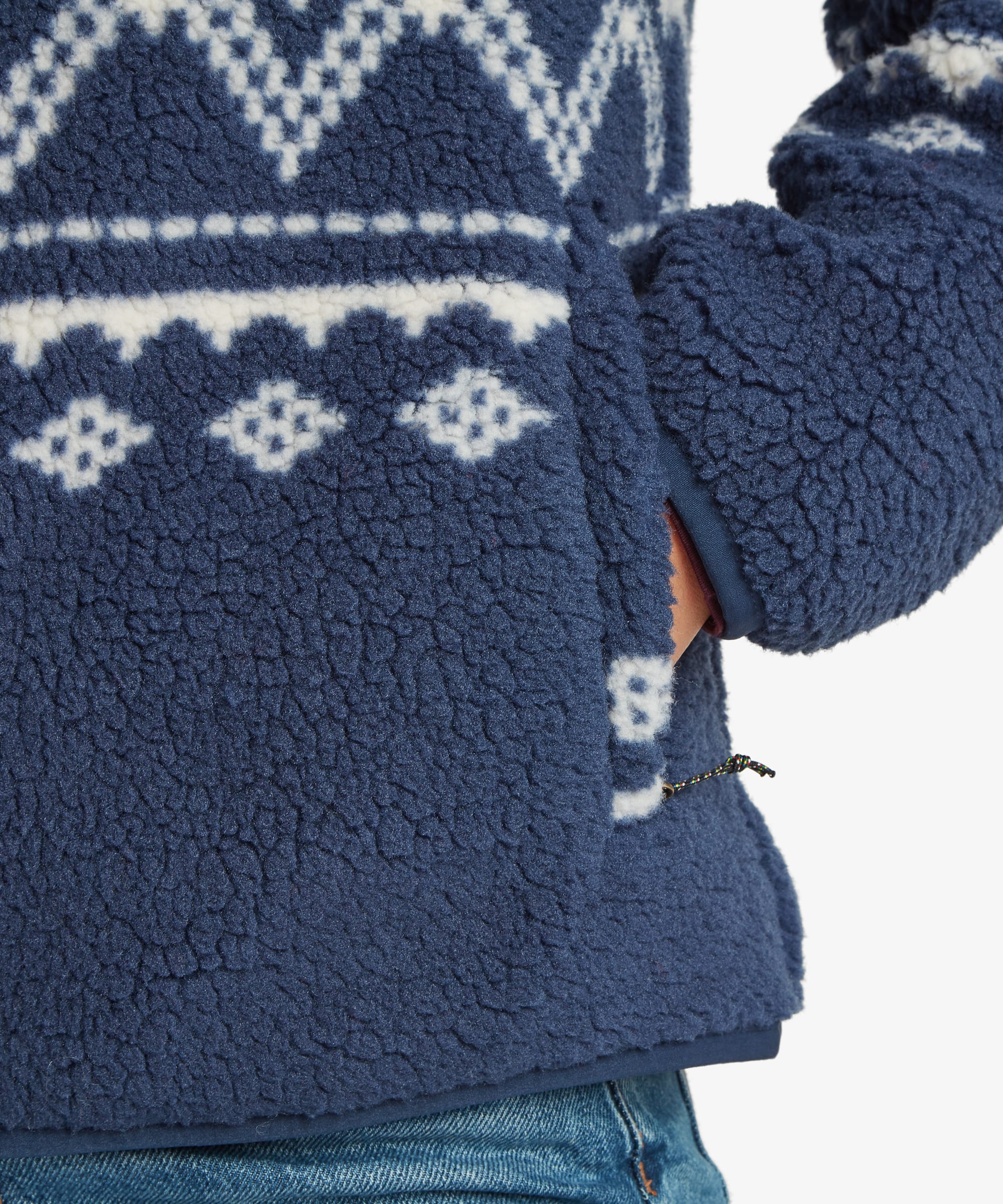 Close-up detail of the sleeve and side pocket of a blue fleece jacket worn by a woman. The image highlights the textured sherpa fabric, geometric pattern, and functional pocket design.