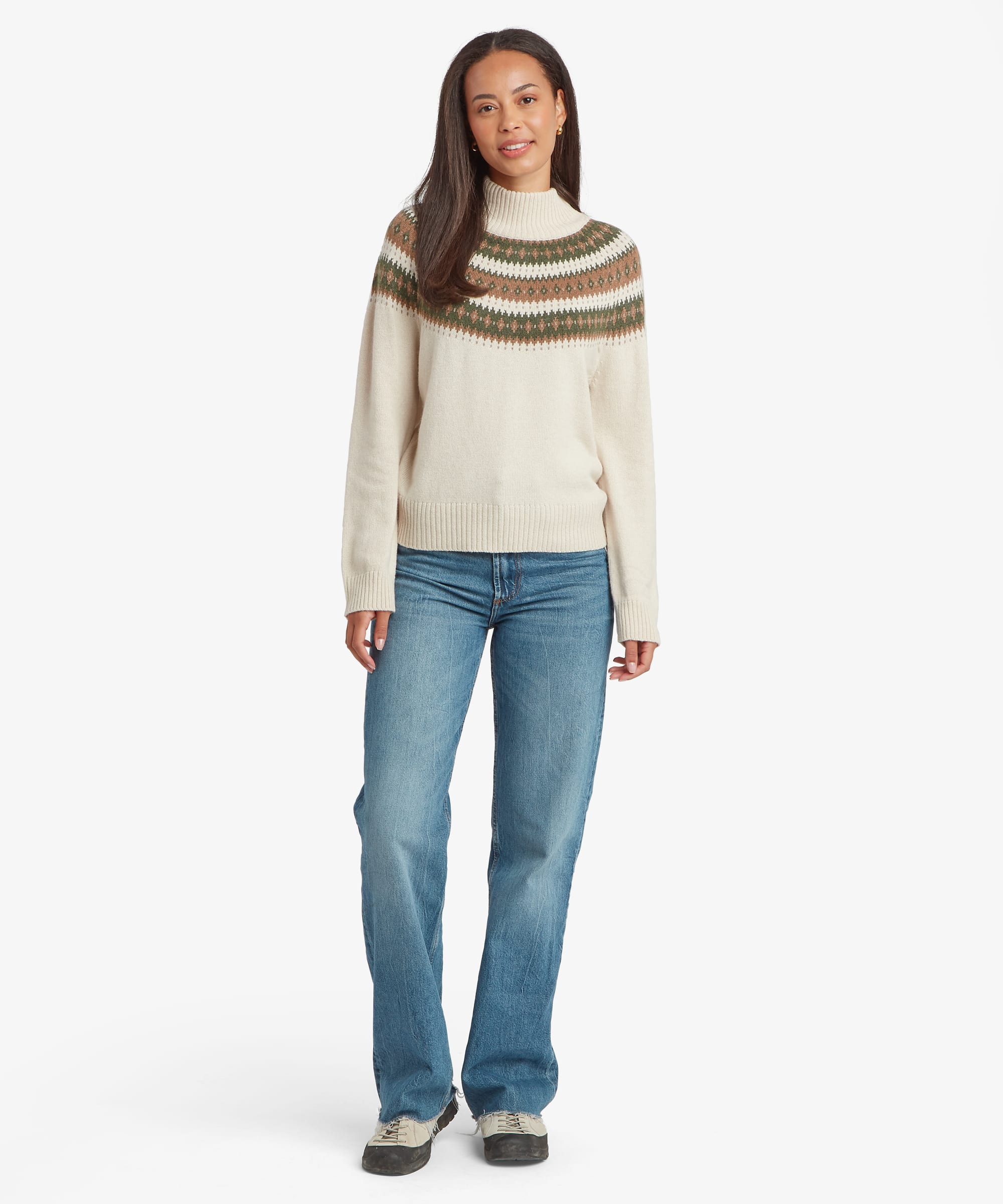 Full-body front view of a woman wearing a cream mock-neck sweater featuring green, brown, and mustard Fair Isle-inspired stripes across the chest and shoulders. The soft knit sweater has long sleeves, ribbed trim, and a comfortable relaxed fit paired with blue jeans.