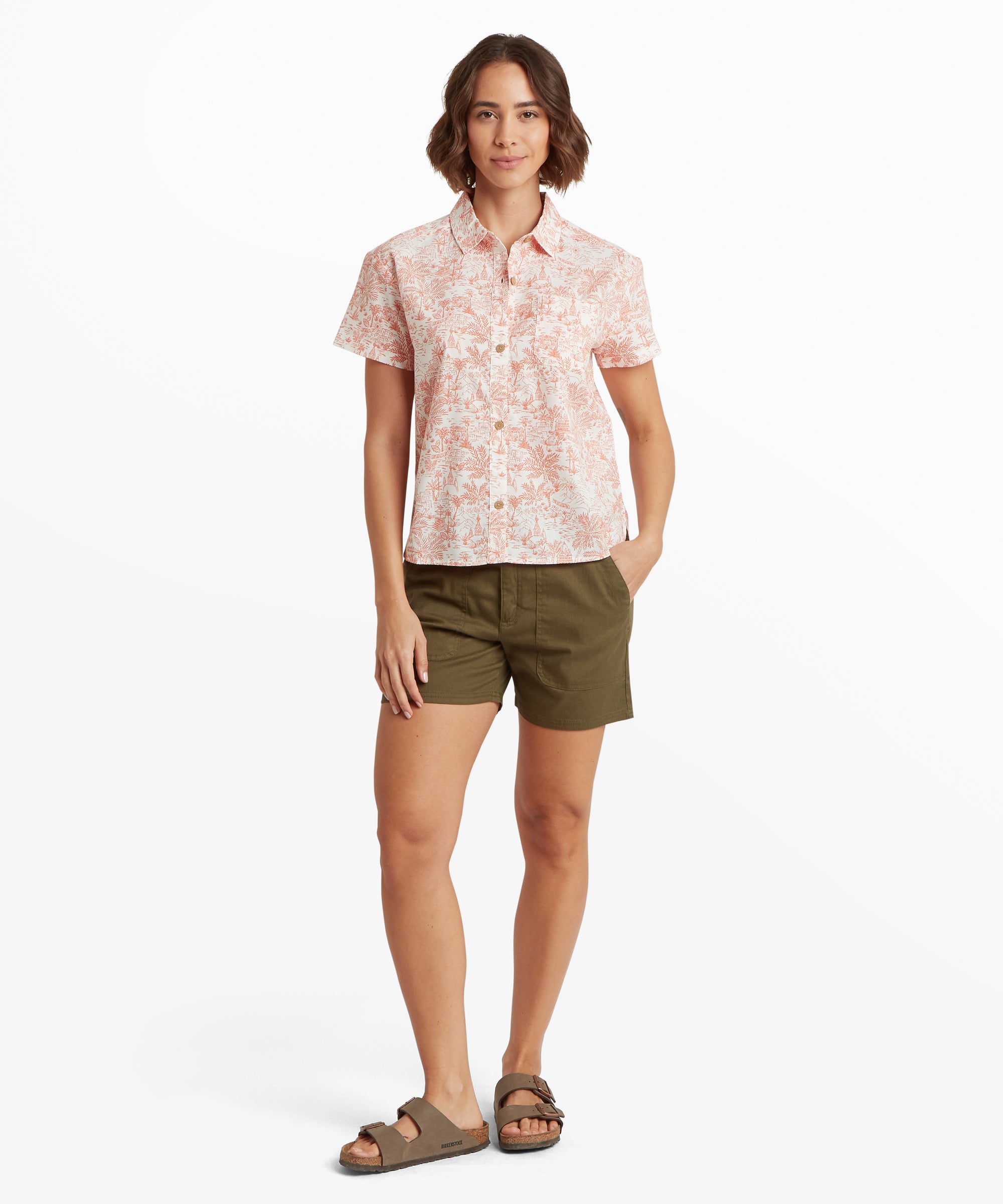 A full-length view of a woman standing in the short-sleeved coral print shirt and olive shorts. The shirt features a loose, boxy silhouette that falls straight through the hips, paired with brown two-strap sandals to emphasize a breezy, travel-ready outfit.