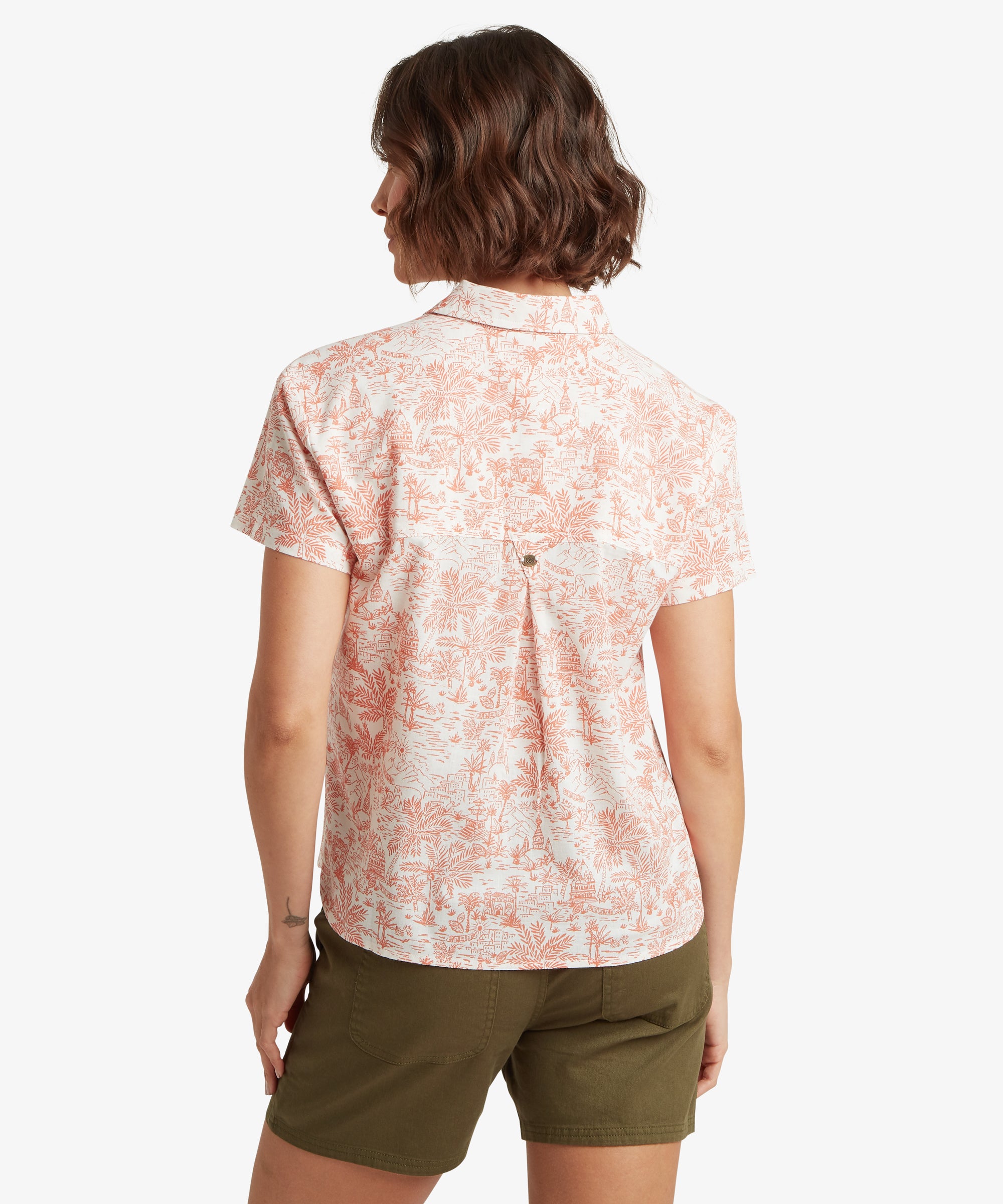 A rear view of a woman wearing the printed shirt, highlighting the straight back yoke and a center box pleat for added roominess. The relaxed silhouette is maintained through the back, showing how the light fabric hangs smoothly over the shoulders. Paired with green shorts.