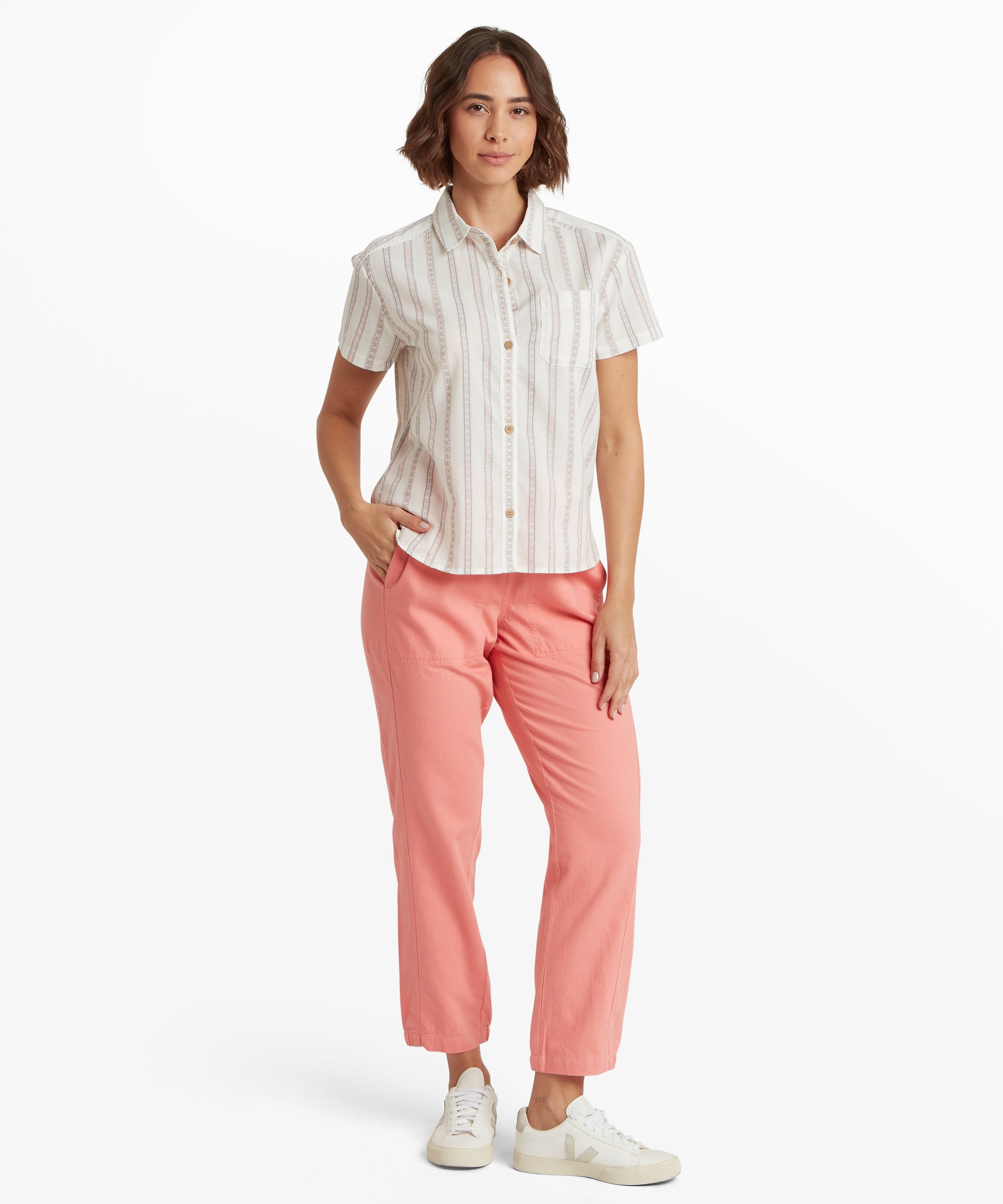 A full-length view of a woman standing in a vertical-striped button-up shirt and pink ankle-length pants. The shirt hangs loosely with a straight hemline that rests at the hip, emphasizing a comfortable, unconstructed silhouette suitable for everyday wear.