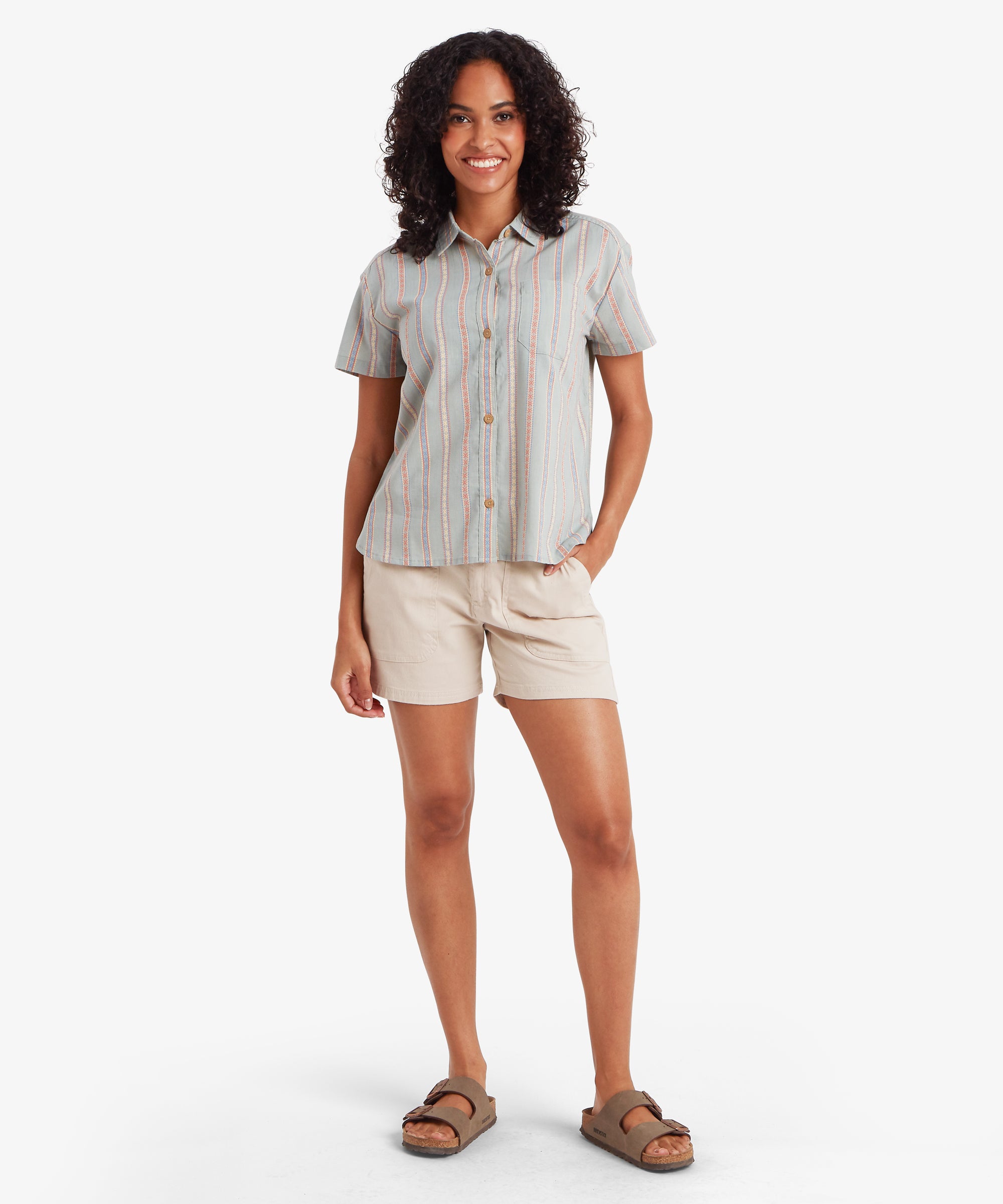 A full-length view of a woman standing in a short-sleeved button-down shirt and light beige shorts. The shirt’s relaxed silhouette is highlighted by the vertical stripe pattern, which creates a long, clean line that pairs easily with the comfortable sandals.