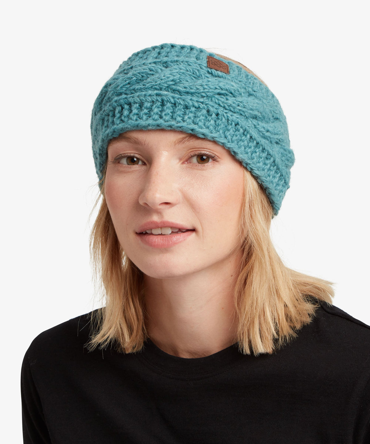 A woman wearing a teal blue chunky knit beanie hat with a small brown brand patch on the side.