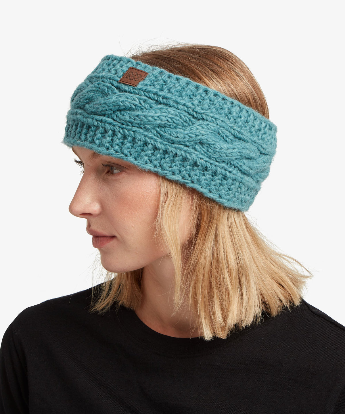 A woman wearing a teal cable-knit headband with a small brown leather logo patch.