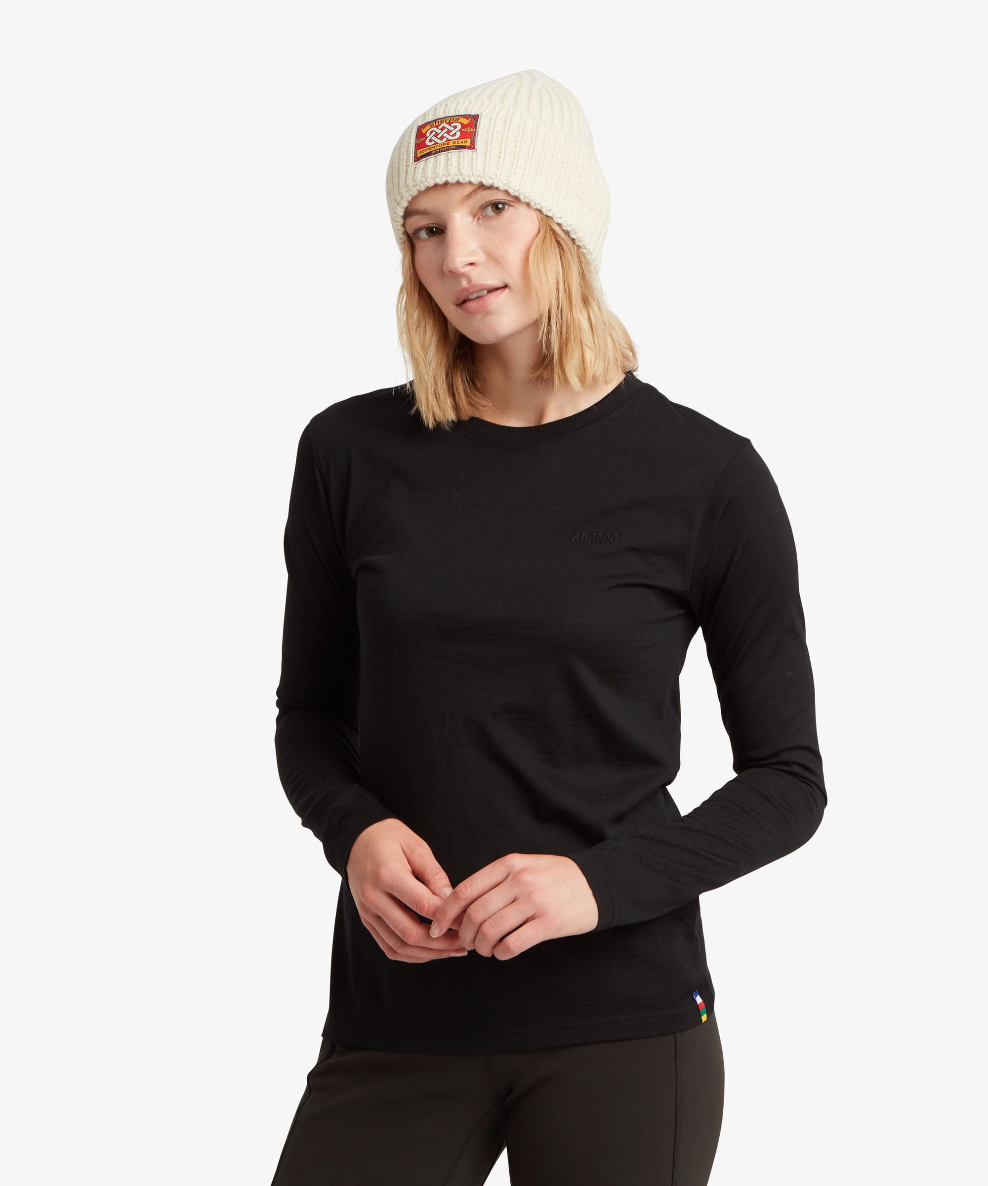 A female model, shown from the front, wearing the Sherpa Adventure Gear women's Nanda Merino Long Sleeve T-Shirt in black. The top has a simple, fitted silhouette with long sleeves and a classic crew neckline. The model is wearing it with black bottoms and a cream-coloured beanie, creating a streamlined cold-weather outfit.