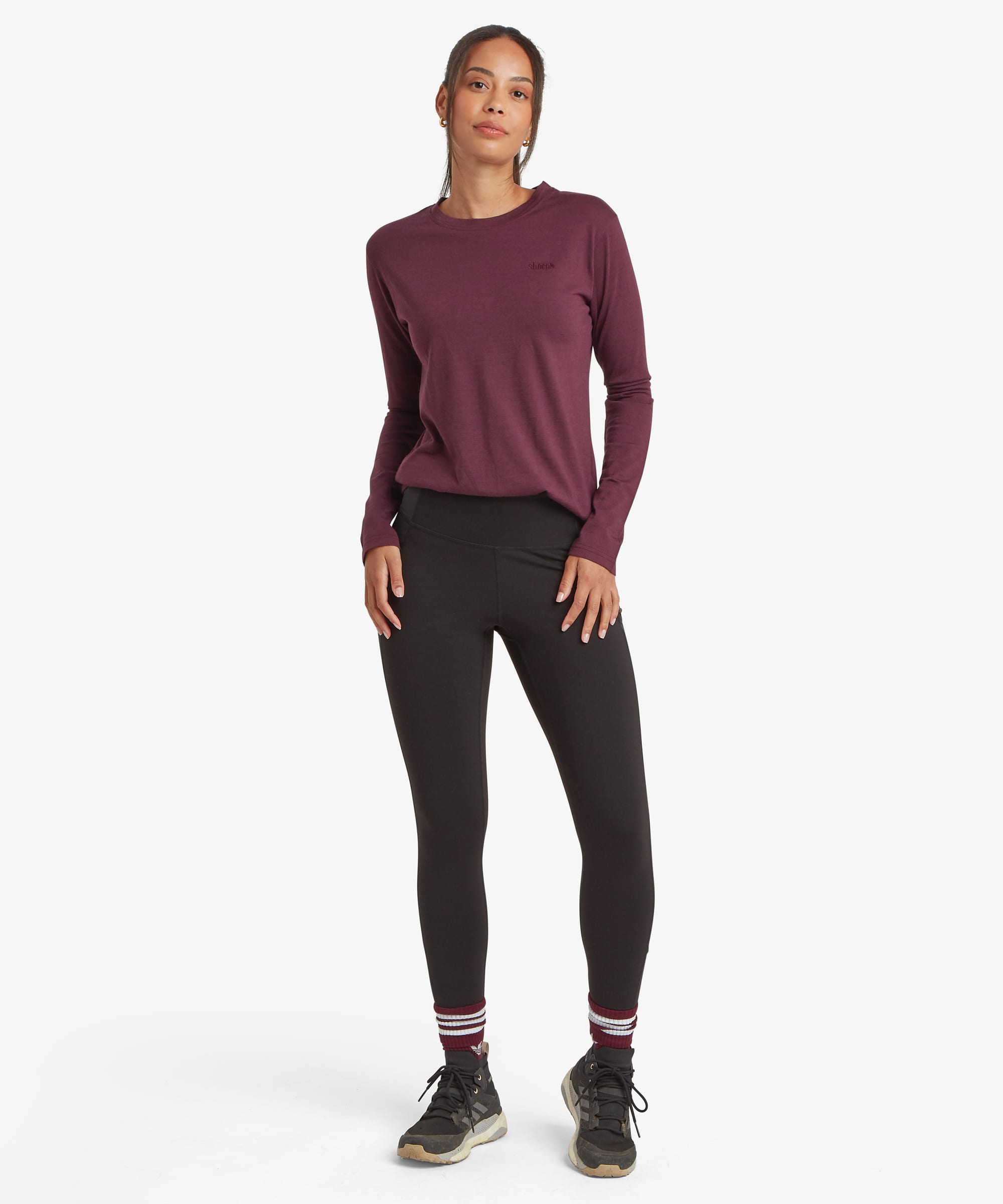 Full-body front view of a woman wearing a burgundy merino long-sleeve T-shirt paired with black leggings. The lightweight performance top features a clean crew neck and fitted silhouette.