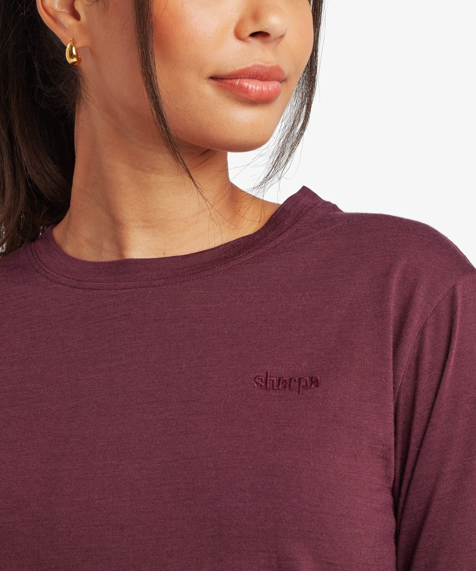 Close-up detail of a woman wearing a burgundy merino long-sleeve T-shirt. The image showcases the soft fabric texture, crew neckline, and subtle embroidered logo on the chest.