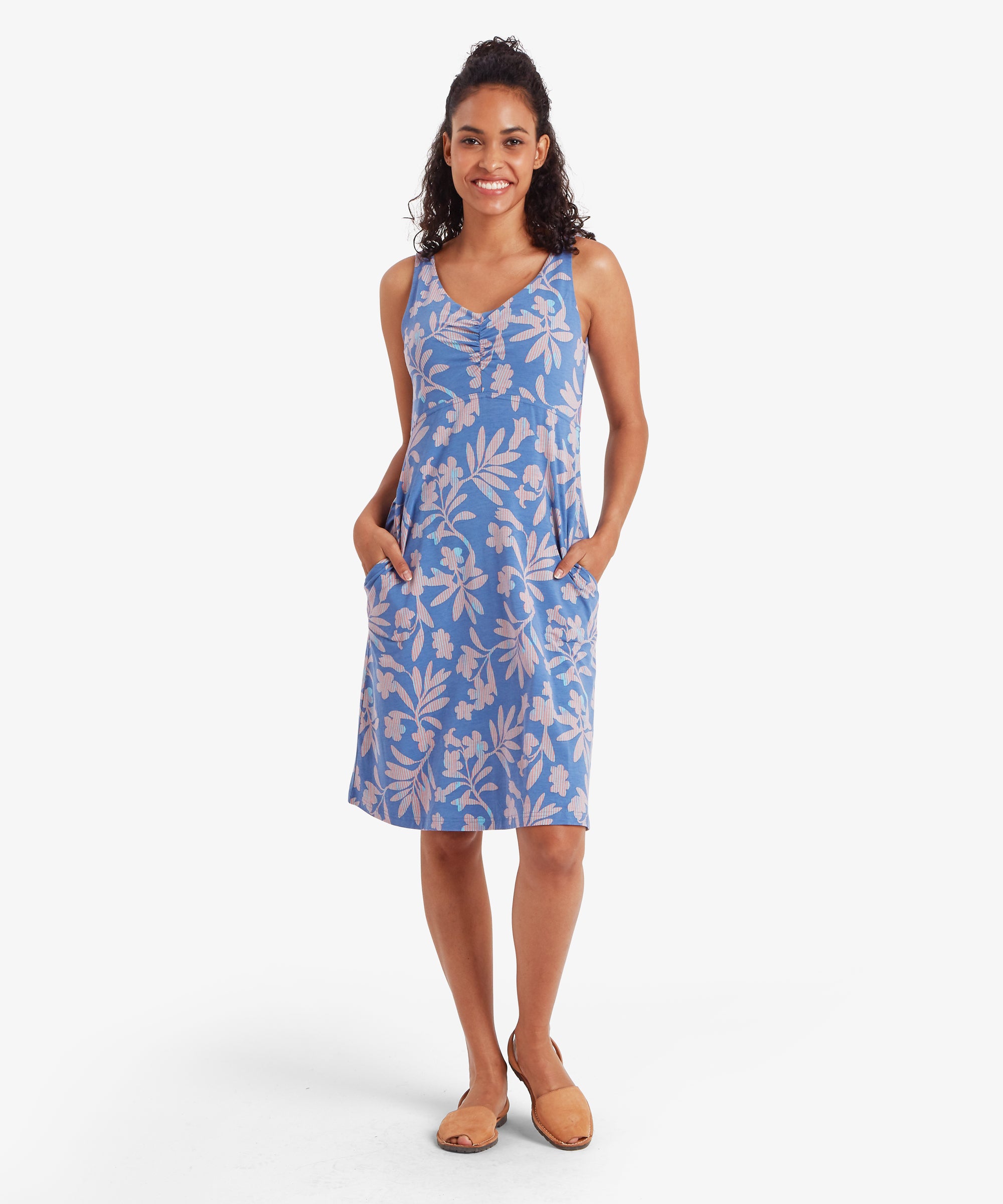 Vorderansicht des blauen Sherpa Neha Empire-Kleides. Die Frau hat beide HŠnde in den Taschen des Kleides.