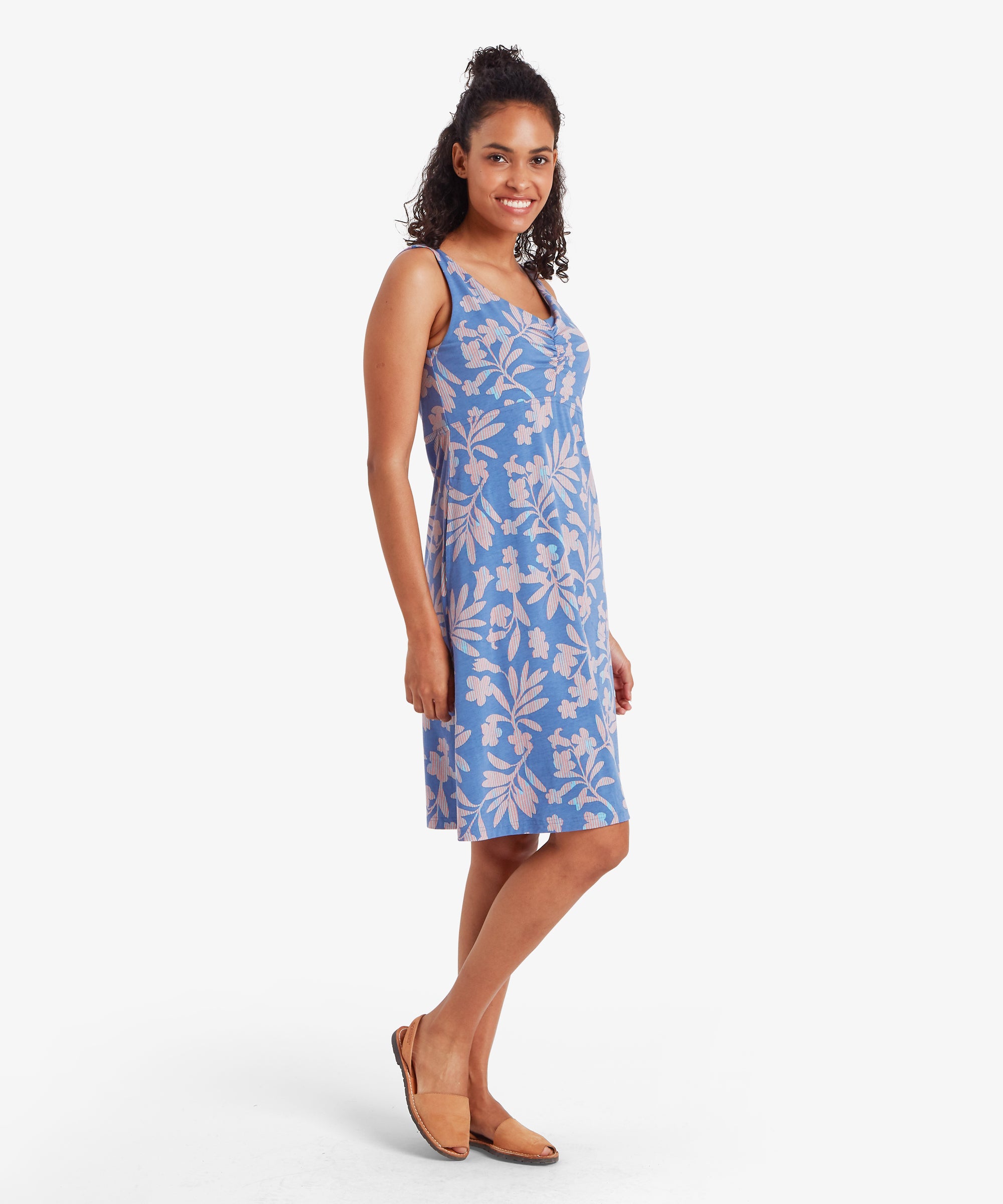 Woman wearing a blue floral dress on a white background.  The woman is standing sideways with one foot slightly raised