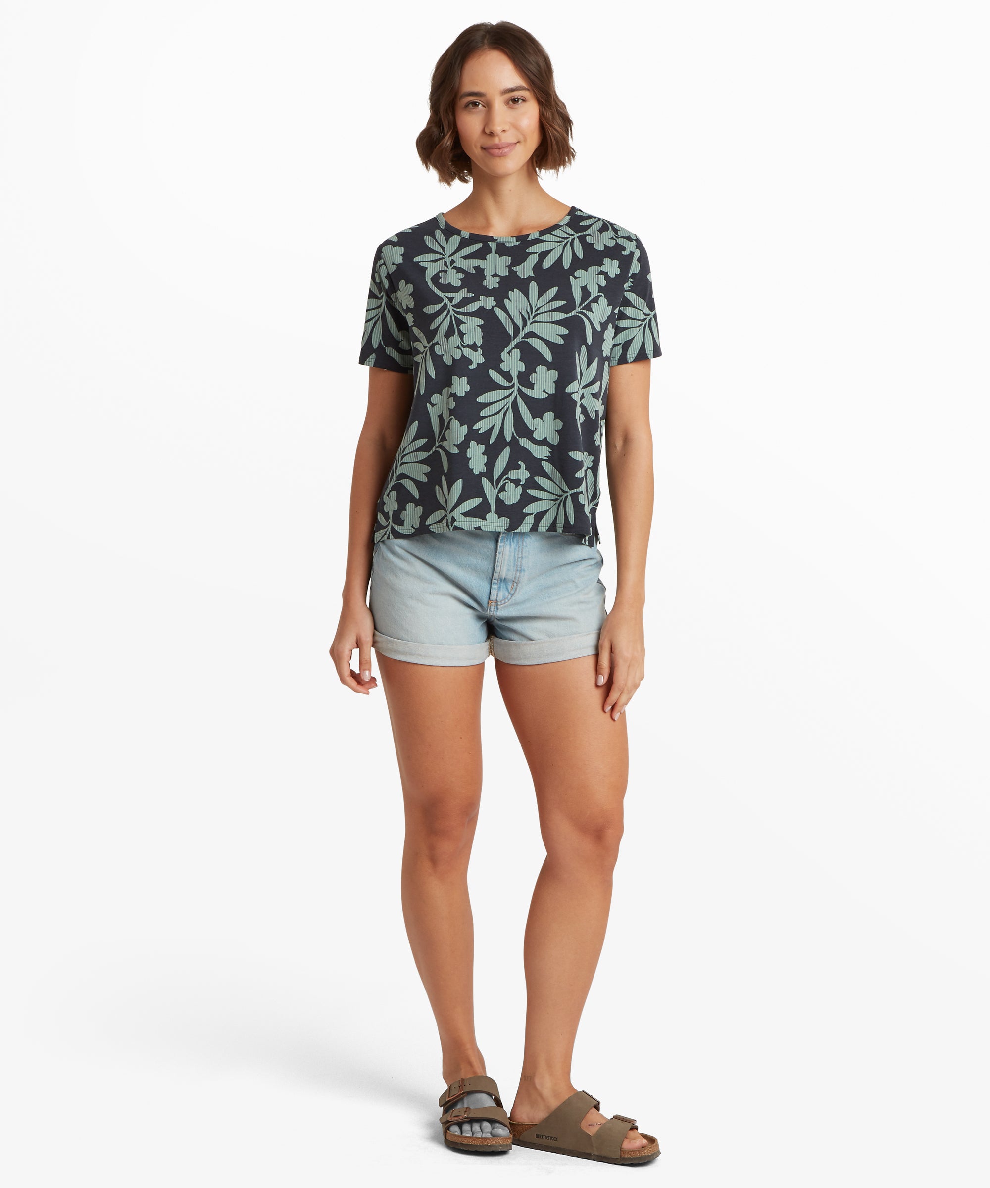 Ganzkšrperansicht eines dunkelblauen T-Shirts mit hellgrŸnem botanischem Print, kombiniert mit hellblauen Jeansshorts mit umgeschlagenem Saum und braunen Sandalen. Das Shirt hat eine lockere Passform und kurze €rmel.