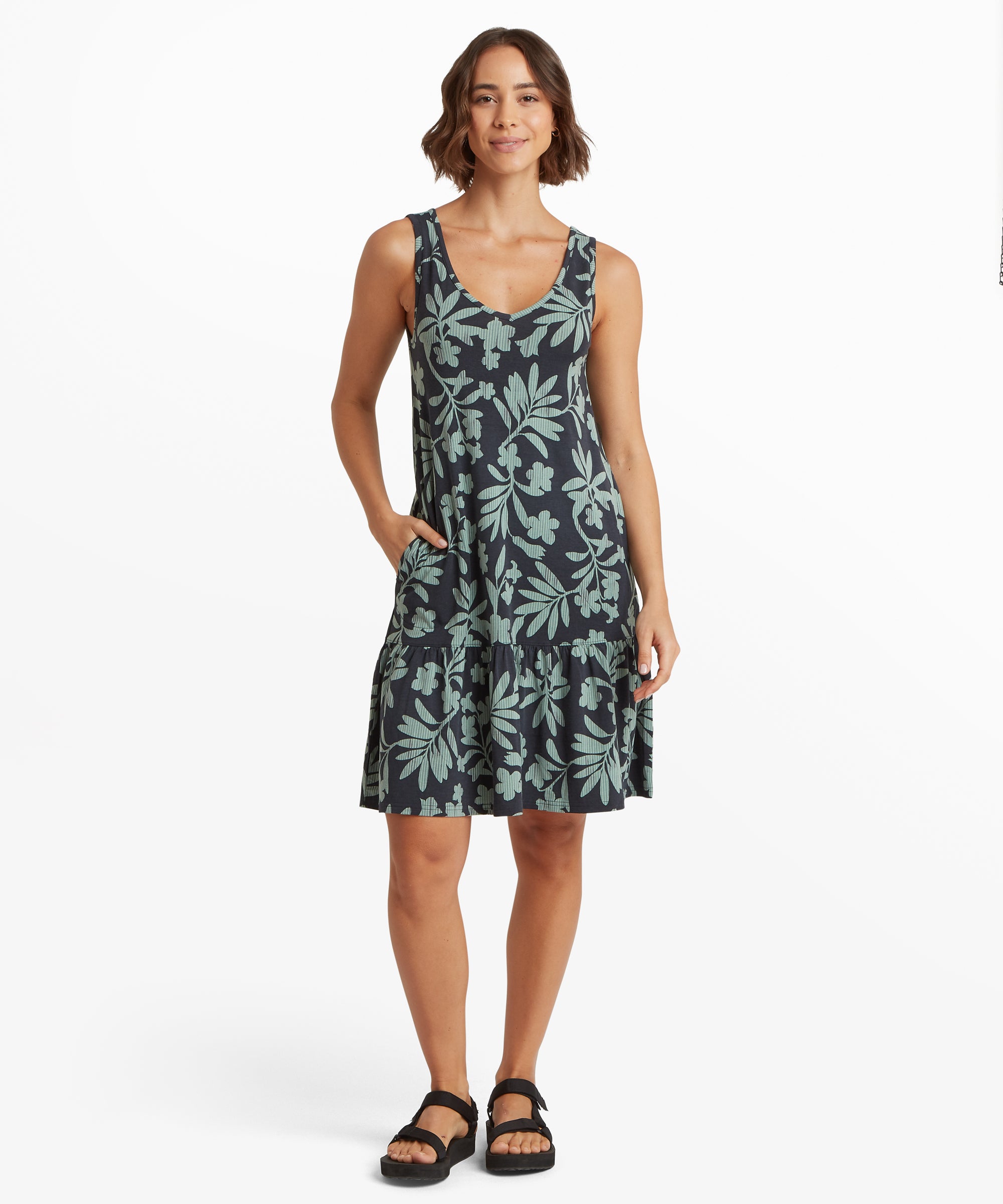 Vorderansicht eines Šrmellosen A-Linien-Kleides mit tŸrkisfarbenem Botanik-Print auf dunklem Hintergrund, U-Ausschnitt und gestuftem RŸschenbesatz am Saum. Eine Hand ist in der Seitentasche.