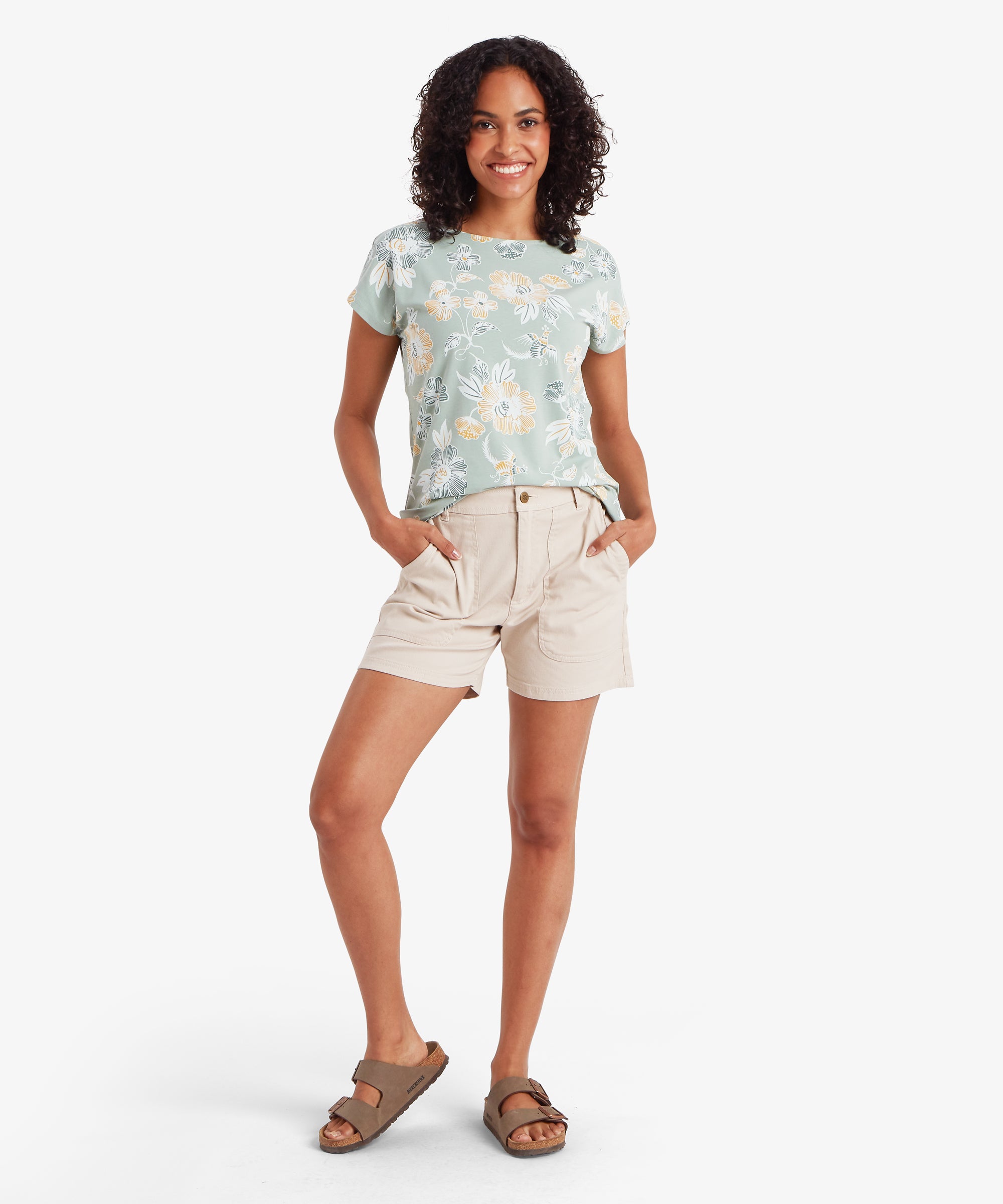 Full‑body view of a light green floral T‑shirt paired with beige shorts and brown sandals, with hands casually in pockets.