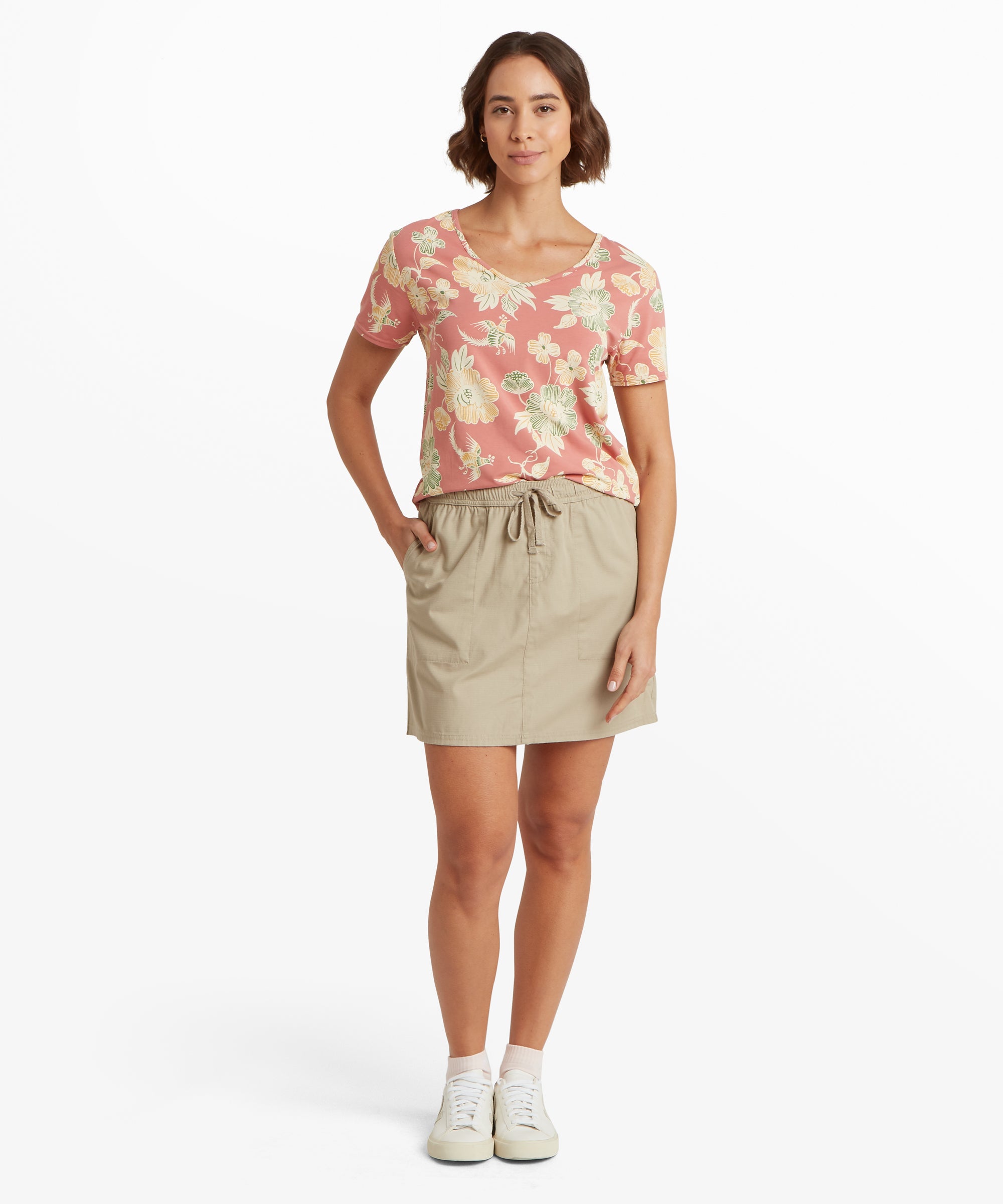 Full‑body view of a pink floral V‑neck tee tucked casually into a beige drawstring skirt, paired with clean white lace‑up sneakers.