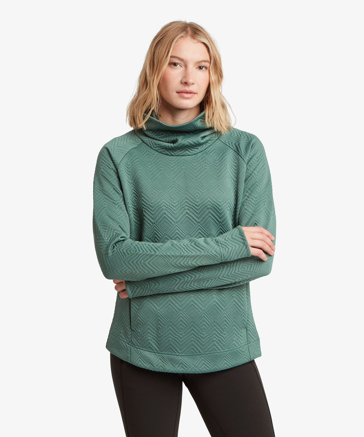 A woman modeling a green textured long-sleeve athletic top with a cowl neck, paired with black leggings.