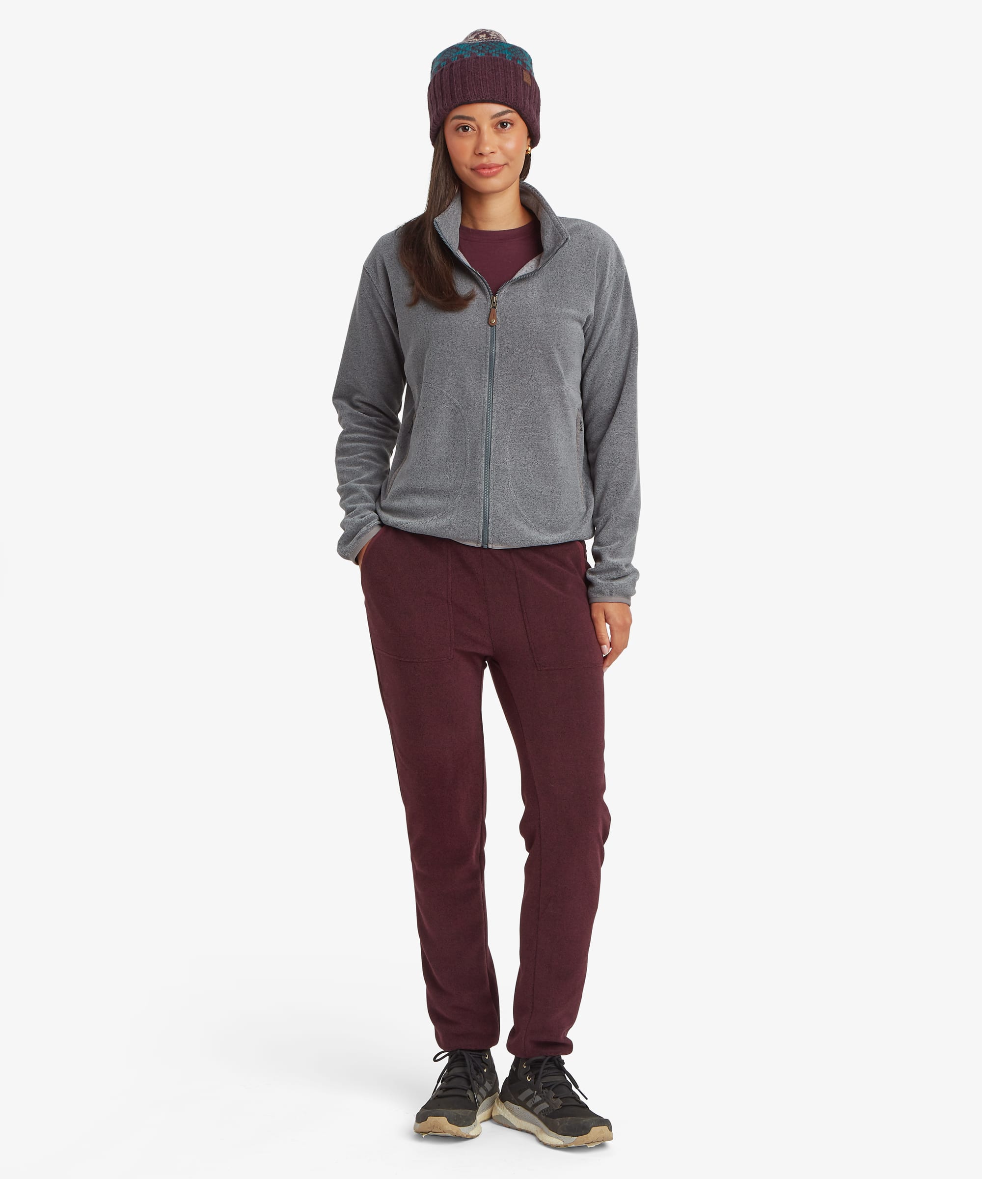 Full-body view of a woman wearing the Sherpa Rolpa Full-Zip Fleece Jacket in grey with burgundy trousers and a knitted beanie. The image highlights the comfortable fit and versatile outdoor styling.