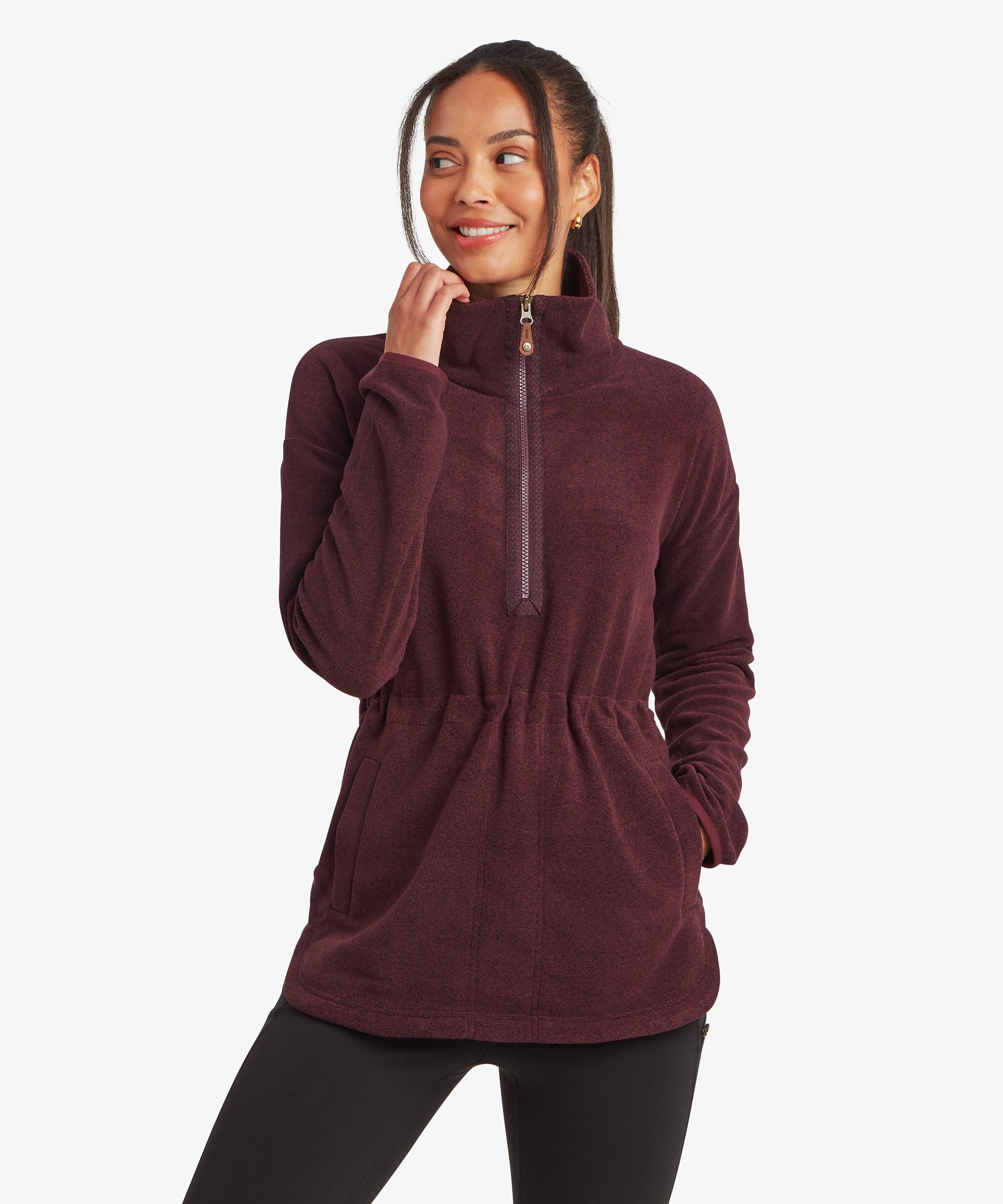 Woman wearing a burgundy Sherpa Rolpa Pop Over fleece with a high funnel neck and adjustable drawcords, standing at a slight angle with one hand raised to the collar. The pullover features soft textured fleece fabric, a kangaroo pocket and a curved hem, shown against a white studio background.
