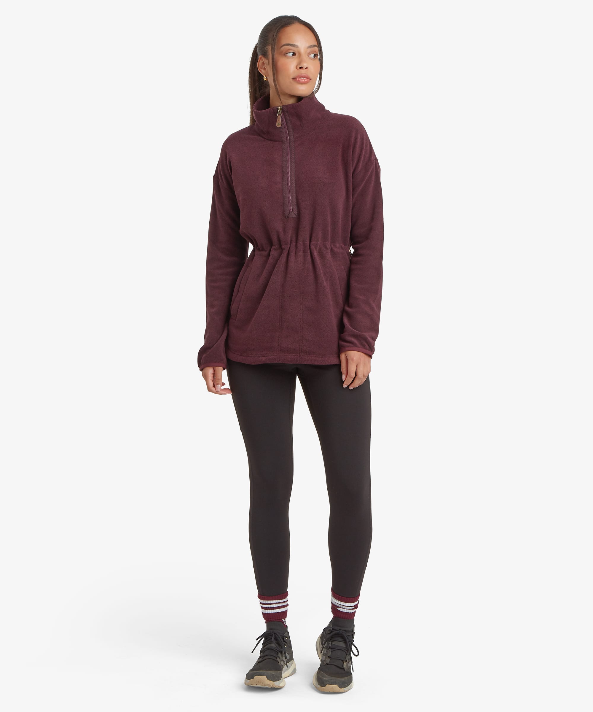 Full-length front view of a woman wearing a burgundy Sherpa Rolpa Pop Over fleece paired with black leggings and trainers. The fleece features a high drawcord neckline, kangaroo pocket and soft textured fabric, highlighting its relaxed fit against a white studio background.