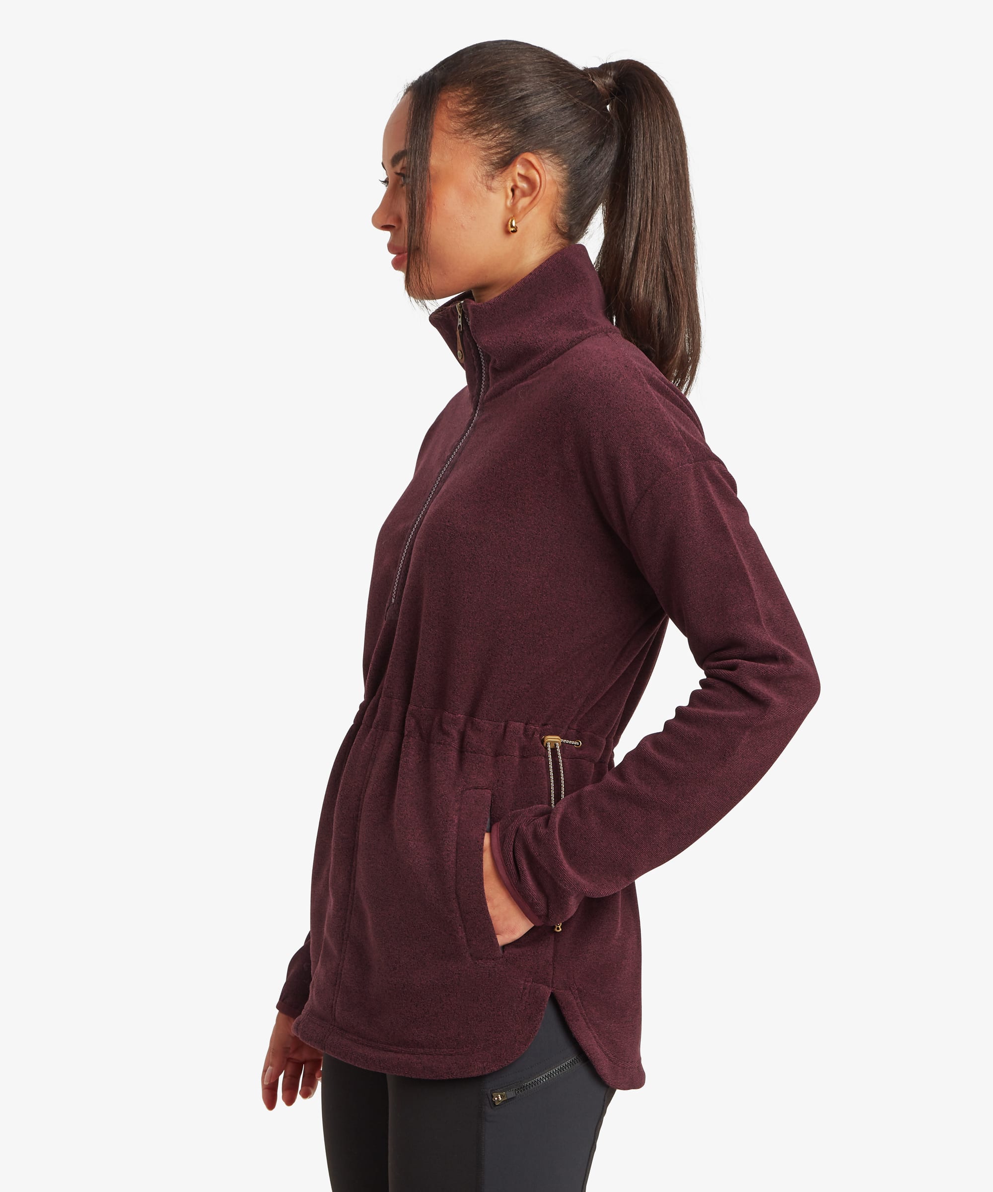 Side profile view of a woman wearing a burgundy Sherpa Rolpa Pop Over fleece. The image highlights the relaxed fit, high funnel neck, side hem vent and kangaroo pocket, shown against a white studio background.