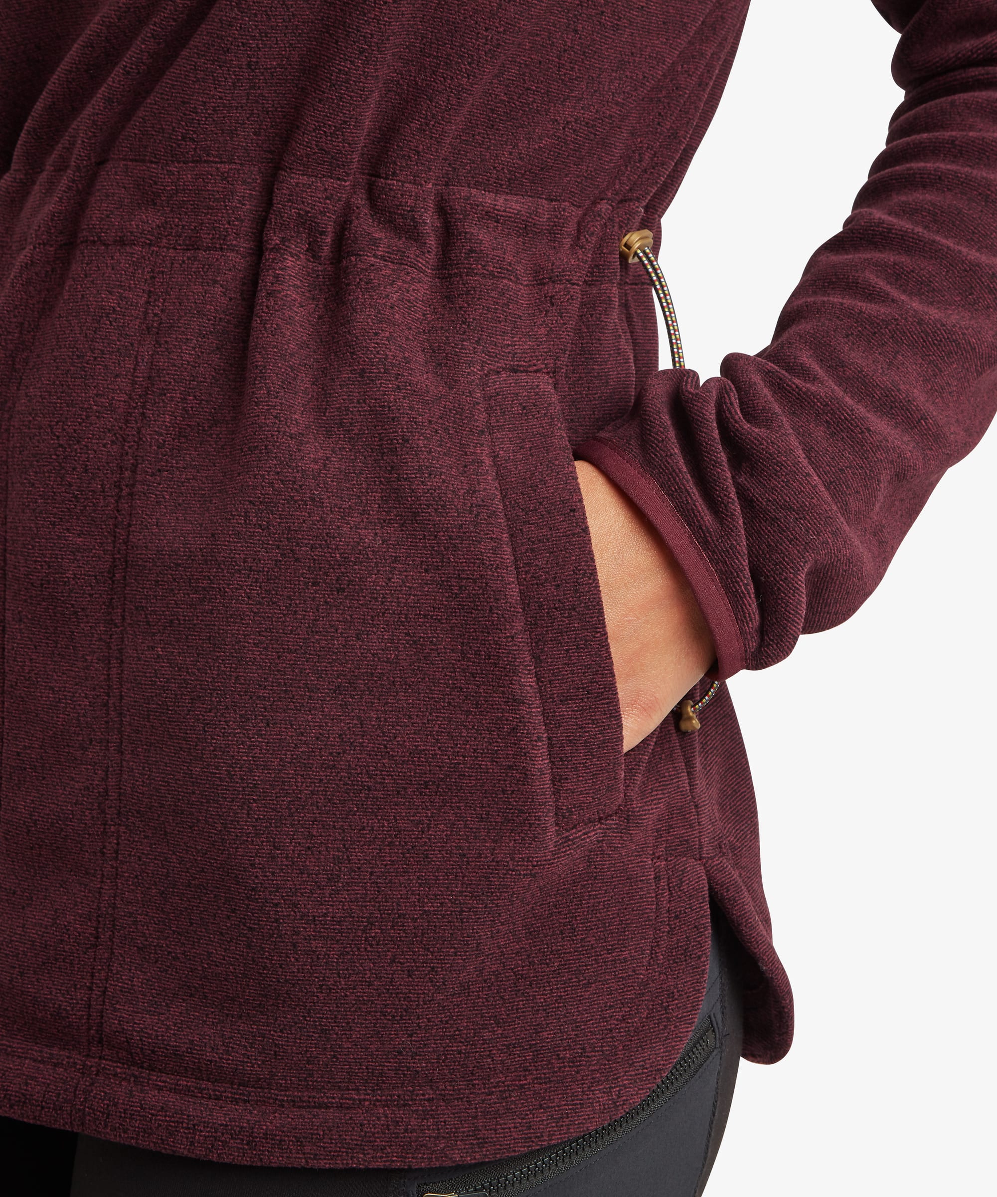 Close-up detail of the burgundy Sherpa Rolpa Pop Over fleece showing the side seam pocket with the model's hand inside. The textured fleece fabric and curved hem are clearly visible.
