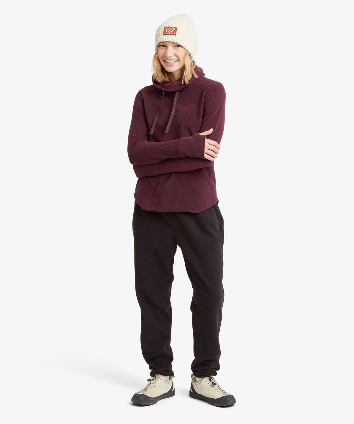 Full-length view of a smiling woman wearing a purple fleece cowl-neck pullover, black pants.