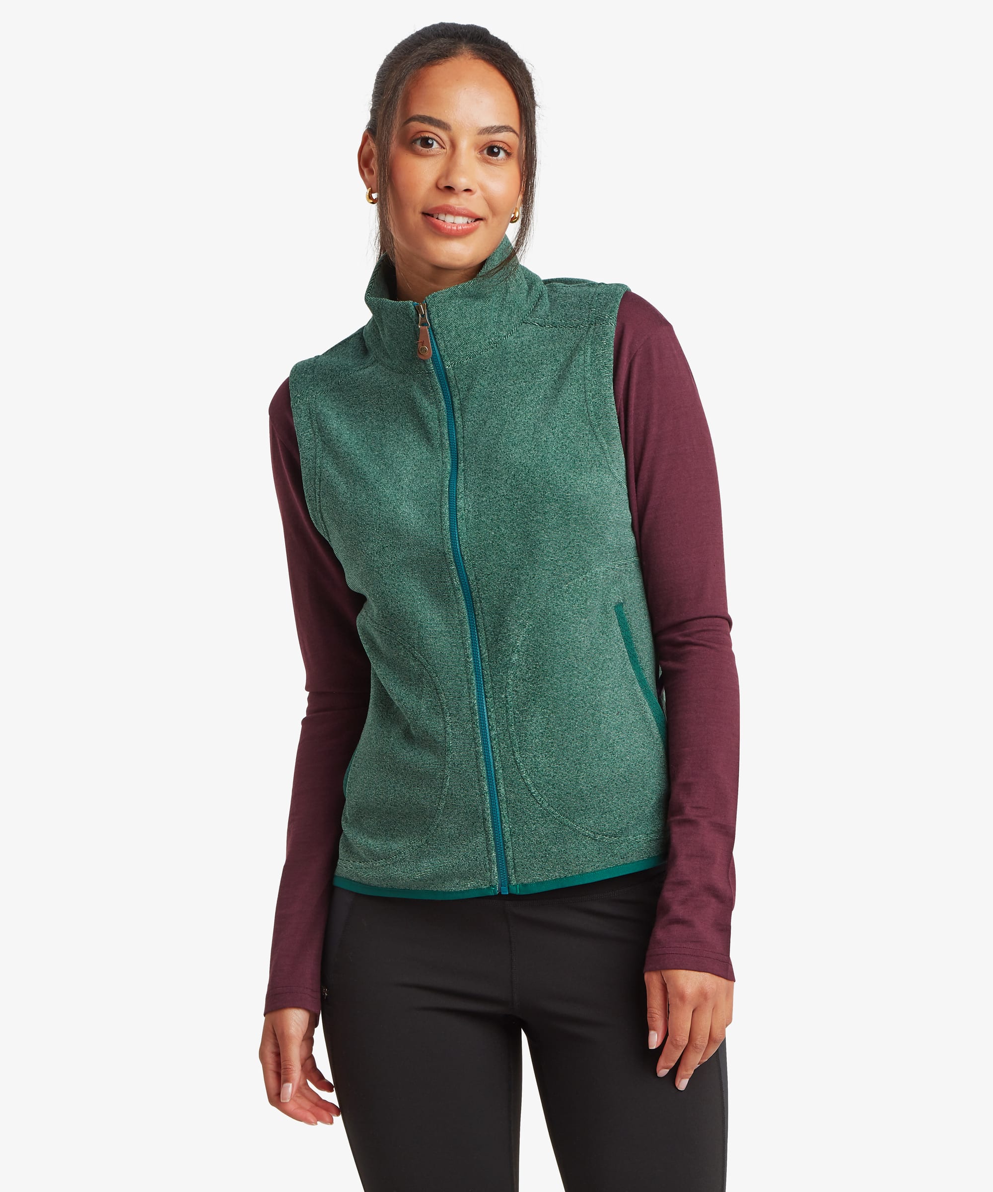Front view of a woman wearing a teal green Sherpa Rolpa fleece vest layered over a burgundy long-sleeve top and black leggings. The sleeveless vest features a full-length zip front, stand collar and soft textured fleece fabric against a white studio background.