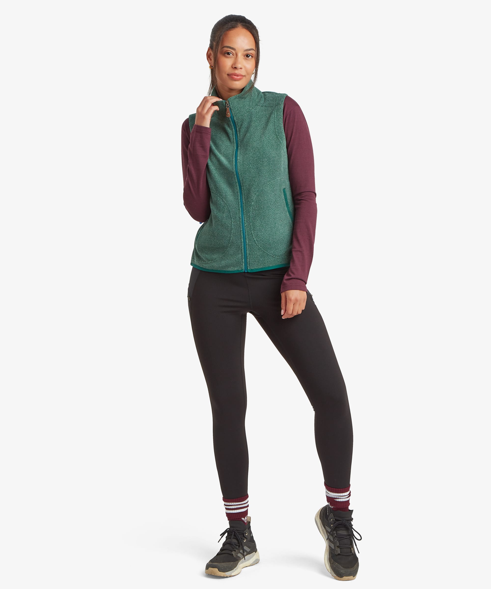 Full-length studio image of a woman wearing a deep green fleece vest layered over a burgundy long-sleeve top and black leggings. She stands facing forward with a relaxed posture, showcasing the vest's full-zip design, high collar, and streamlined fit.