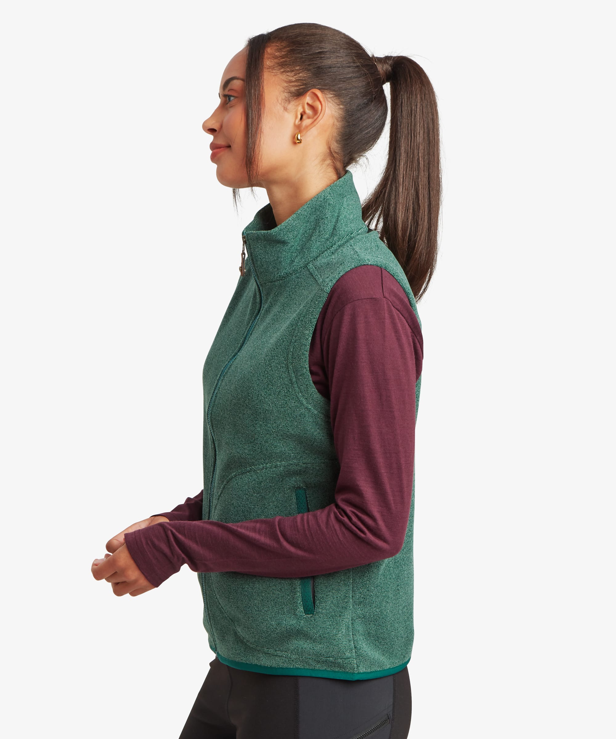 Side-profile studio view of a woman wearing a deep green fleece vest over a burgundy long-sleeve top. The image highlights the vest's sleeveless design, stand collar, and fitted silhouette.