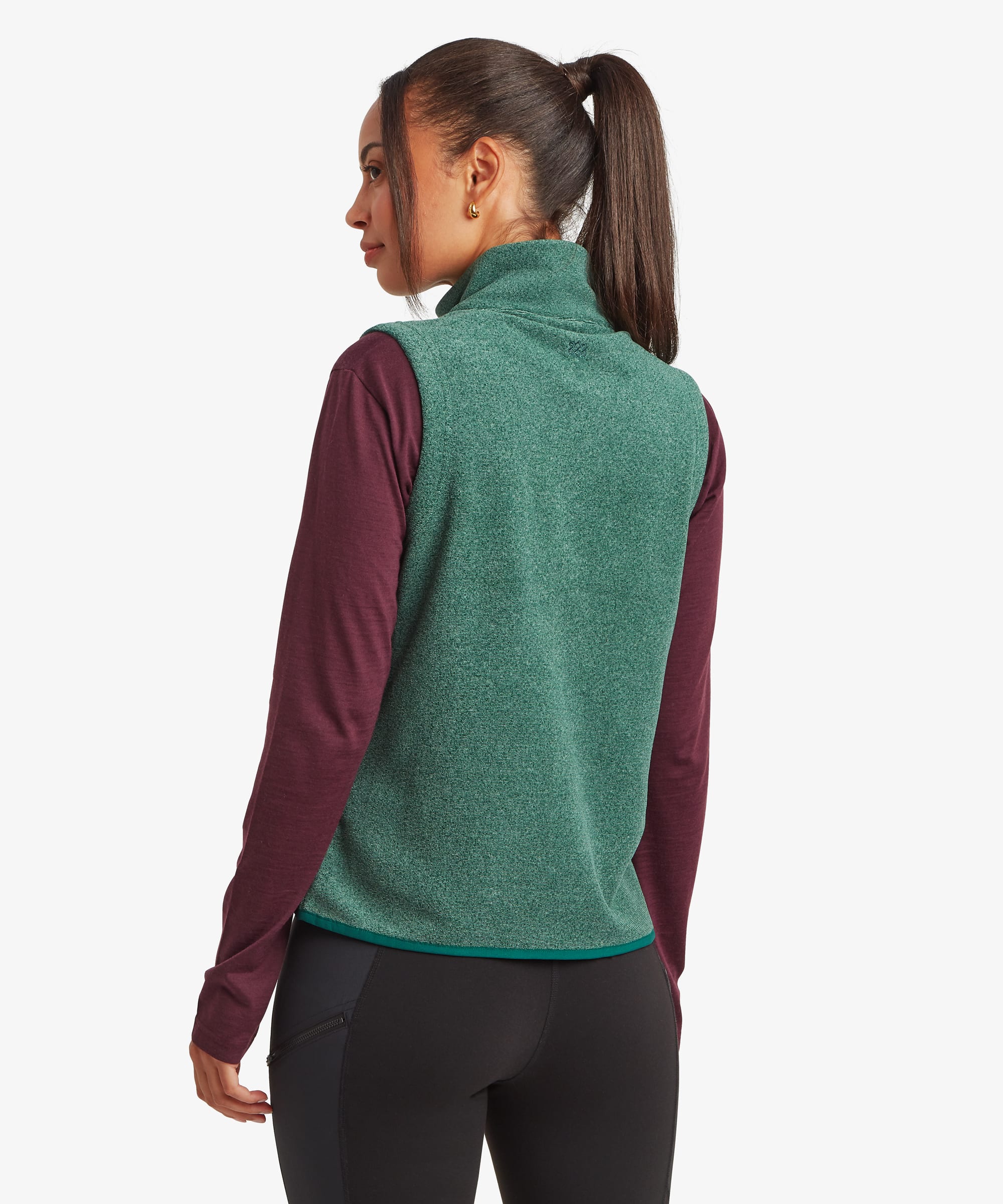 Rear studio view of a woman wearing a deep green fleece vest layered over a burgundy long-sleeve top and black leggings. The image showcases the clean back panel and overall fit of the vest.