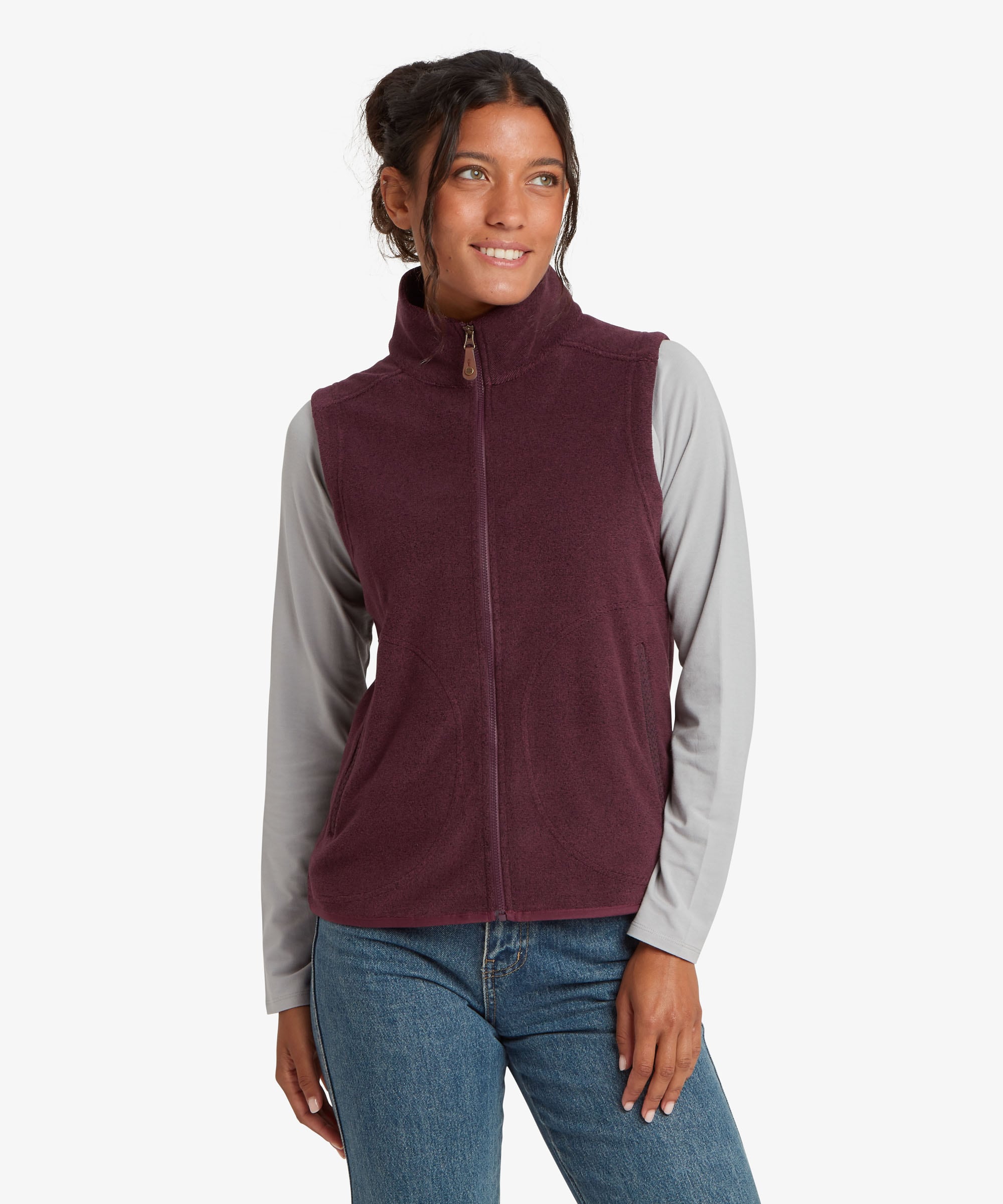 A woman wearing a sleeveless zip-front fleece vest in deep burgundy over a light gray long-sleeve shirt and blue jeans, photographed against a clean white studio background.