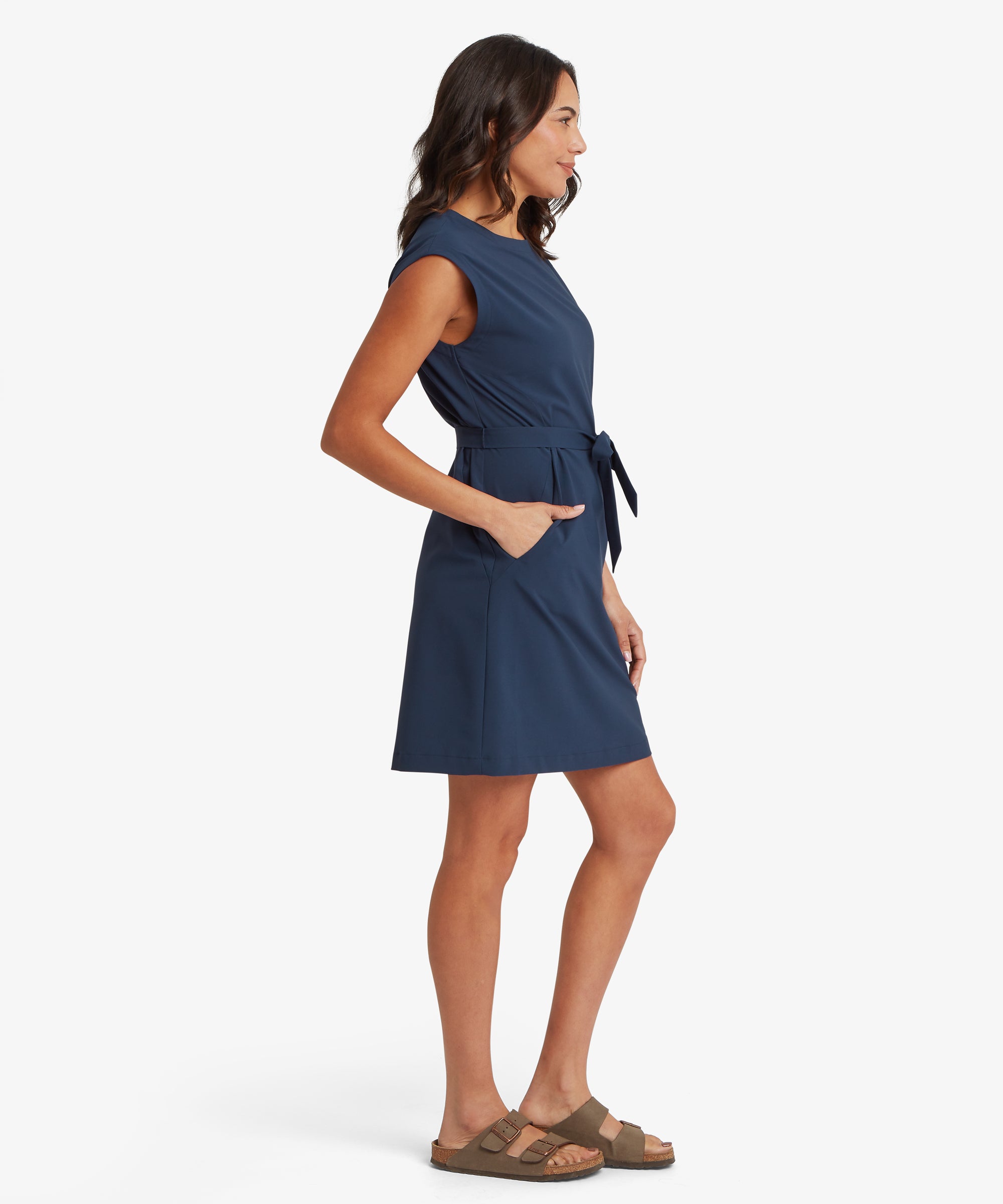 Side view of a woman wearing a navy sleeveless dress with a round neckline and tie waist, showing the gentle drape through the skirt and a relaxed fit through the hips. Her hand is placed in the pocket and her posture is casual.