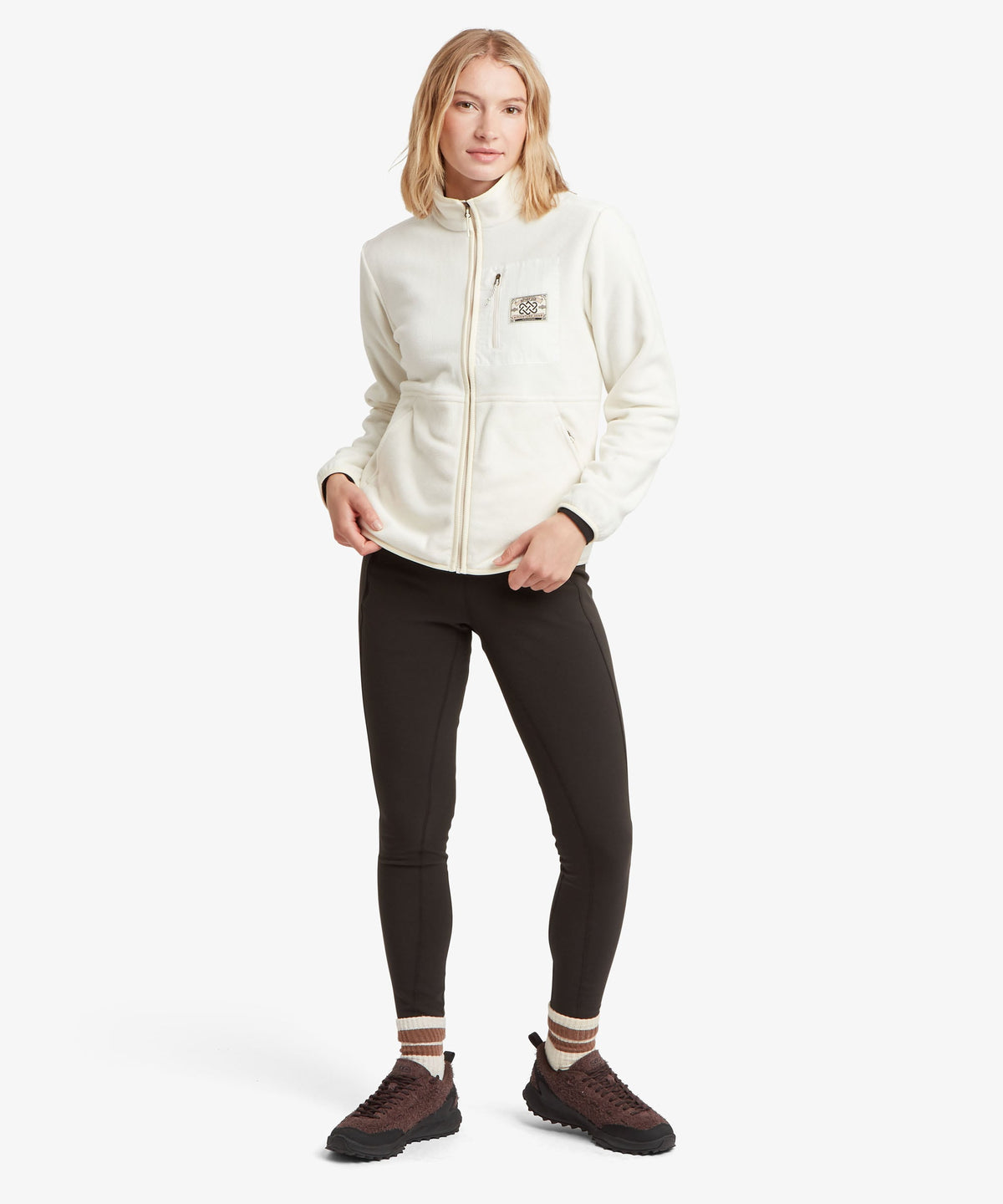 A female model wearing the Sherpa Adventure Gear white Sanu Full-Zip fleece with black trousers.