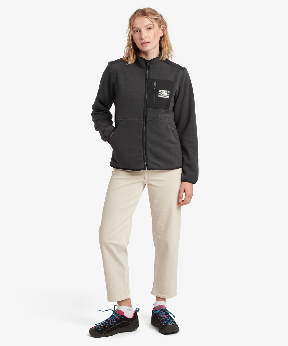 A female model wearing the Sherpa Adventure Gear black Sanu Full-Zip fleece with cream trousers.