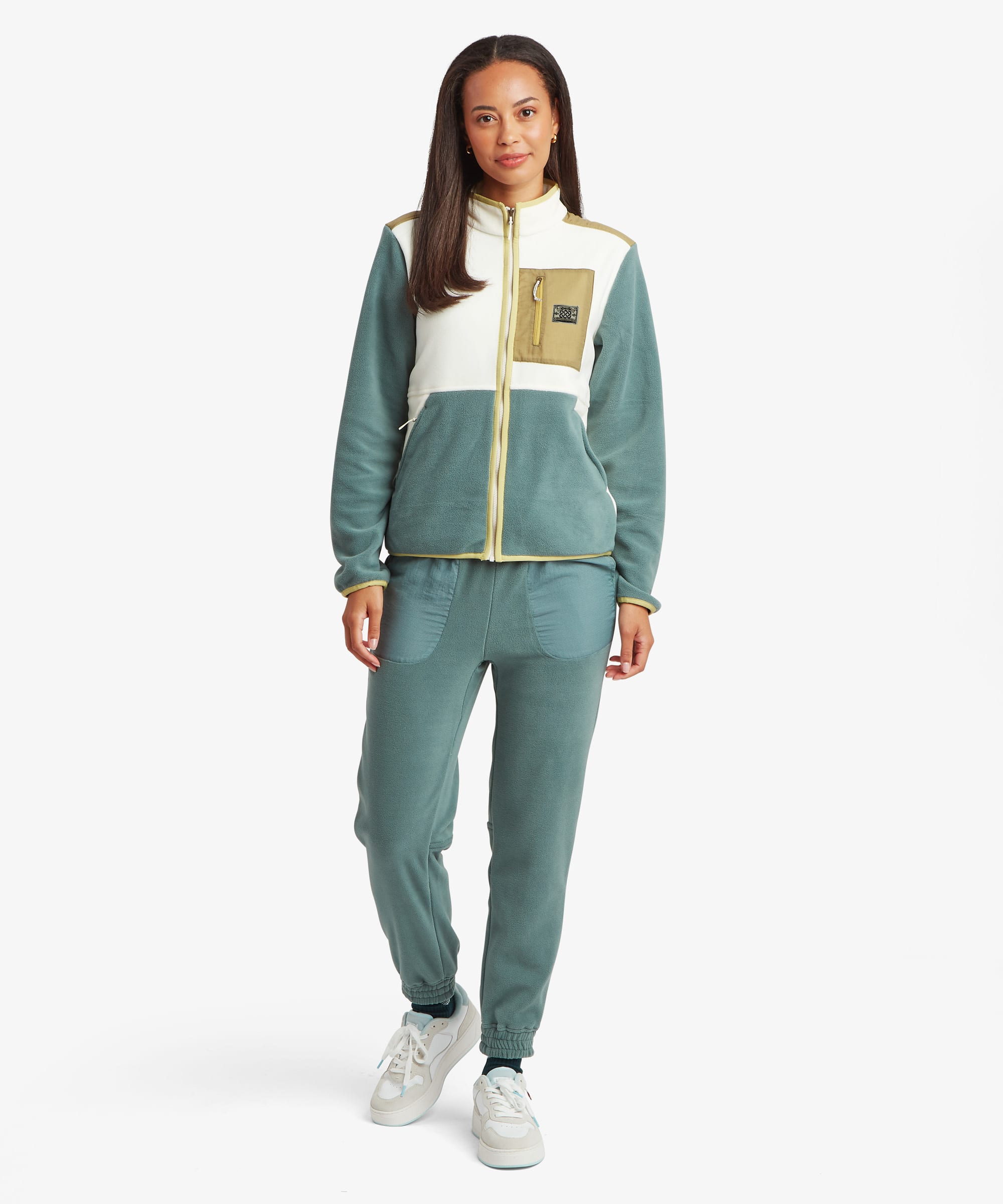Full-length studio image of a woman wearing a colour-block fleece jacket with cream upper panels, green sleeves and lower body, and a tan chest pocket. Styled with coordinating green trousers and white trainers, the image highlights the jacket's overall silhouette.