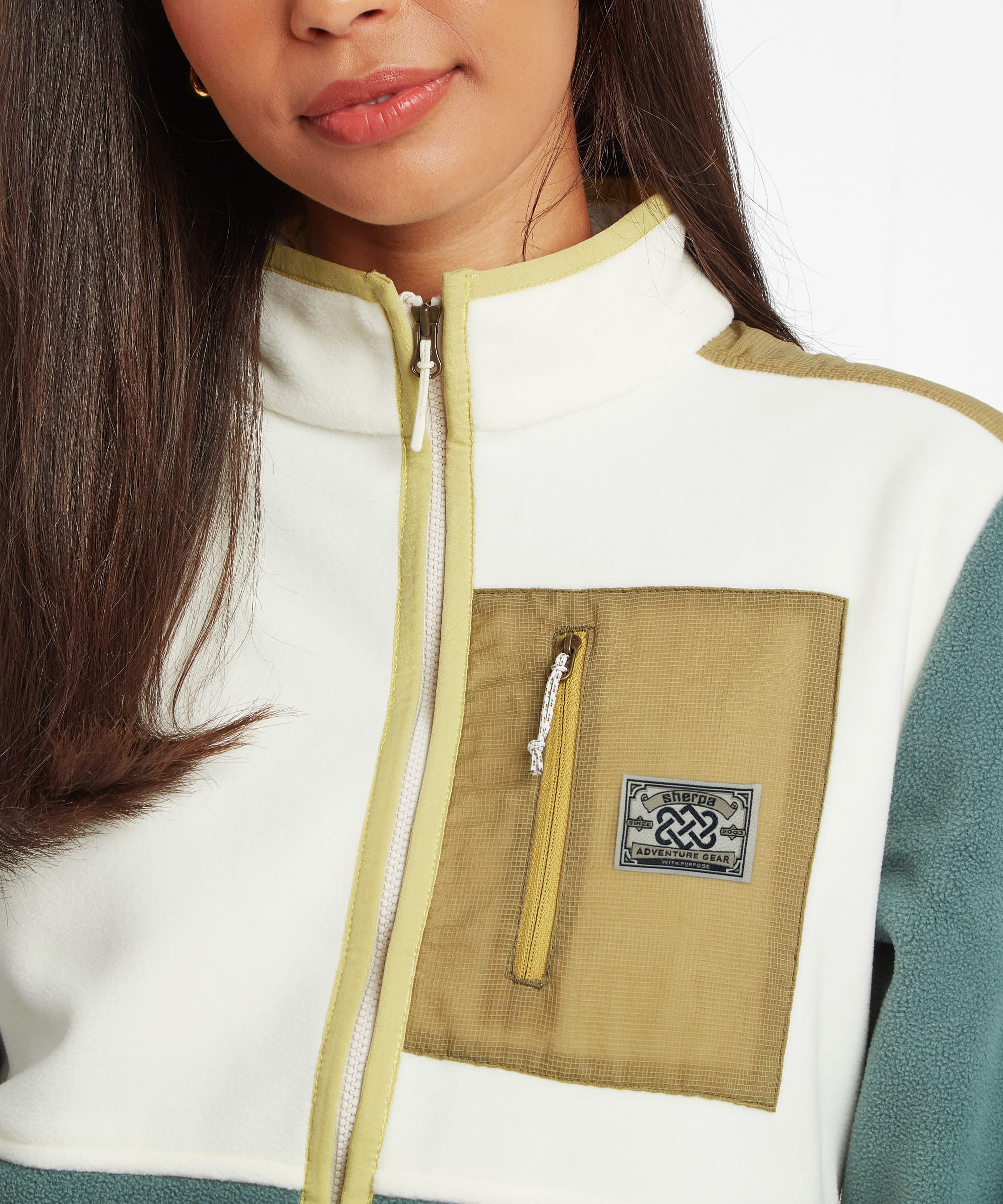 Close-up portrait of a woman wearing a cream and green fleece jacket with a tan utility chest pocket. The image focuses on the high collar, full zip closure, pocket detail, and colour-block design.