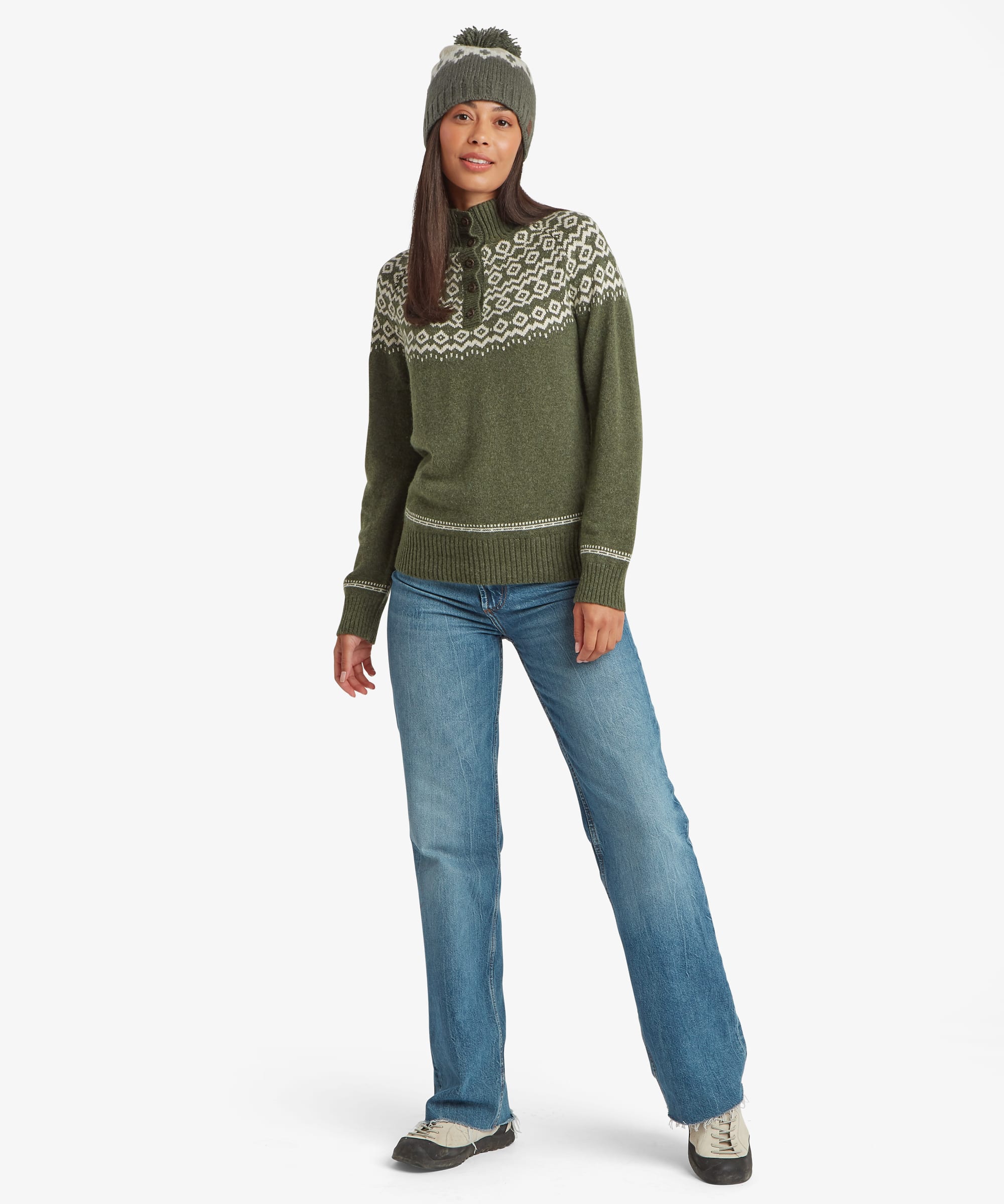 Full-length studio image of a woman wearing an olive-green patterned knit sweater paired with blue jeans and a pom-pom beanie. The image highlights the sweater's classic crew neckline and decorative yoke design.