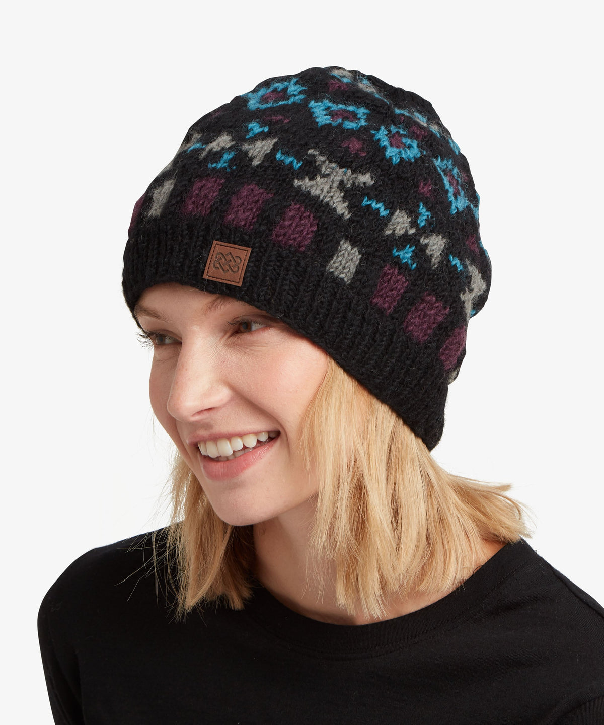 A smiling blonde woman wearing a black knit beanie with a colorful geometric pattern in blue.